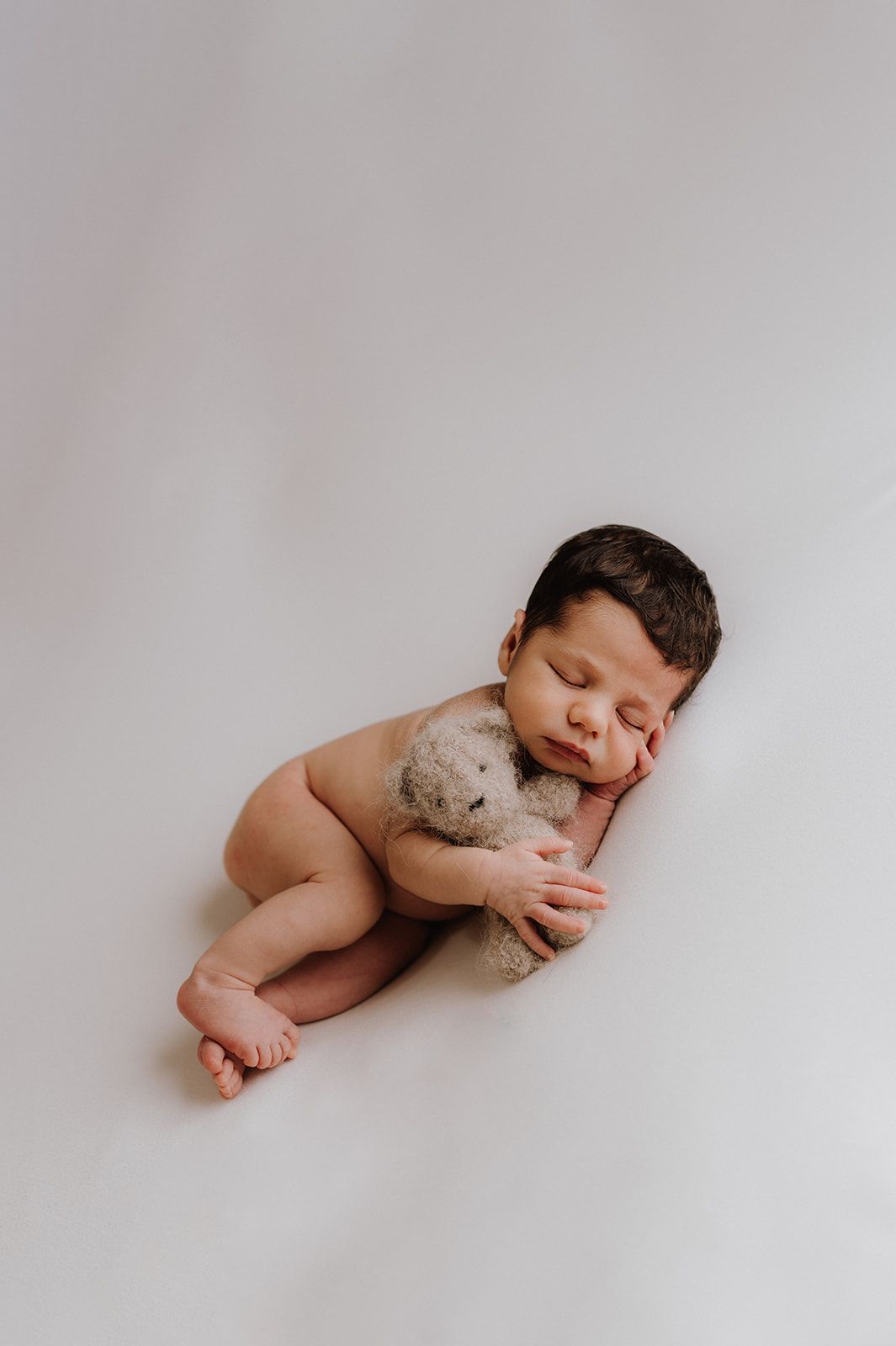Peaceful newborn baby sleeping curled up with a tiny teddy bear on a white background.