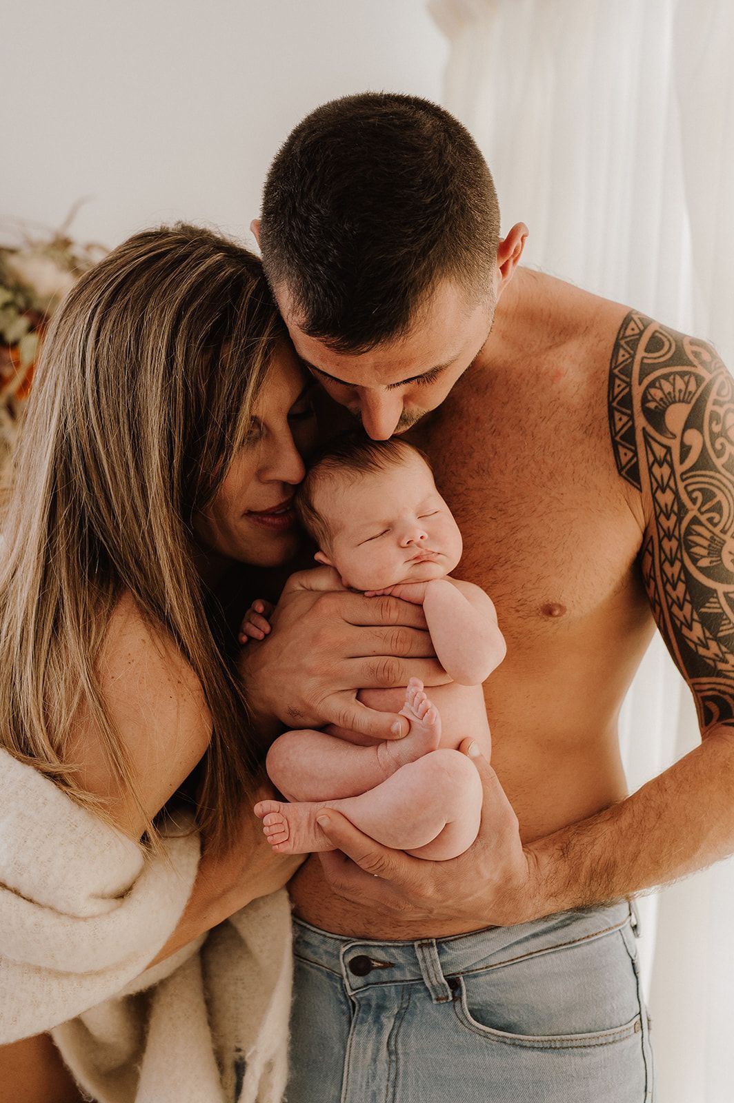 Tattooed shirtless father and mother lovingly hold their sleeping newborn baby in a bright studio.