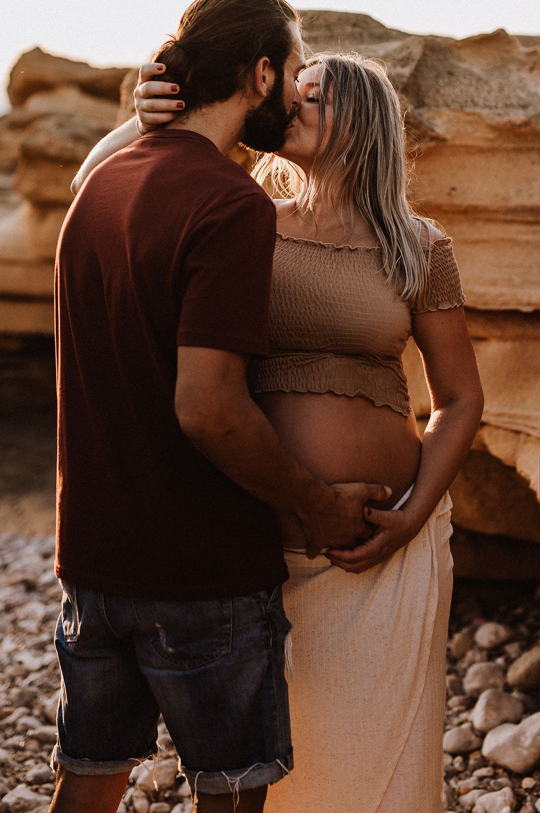 Couple sharing a tender kiss while cradling pregnant belly outdoors on rocky beach at golden hour.