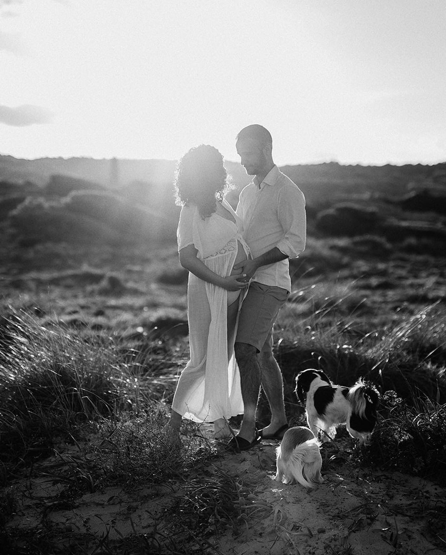 Black and white photo of a couple embracing at sunset in a field with their small dog beside them.