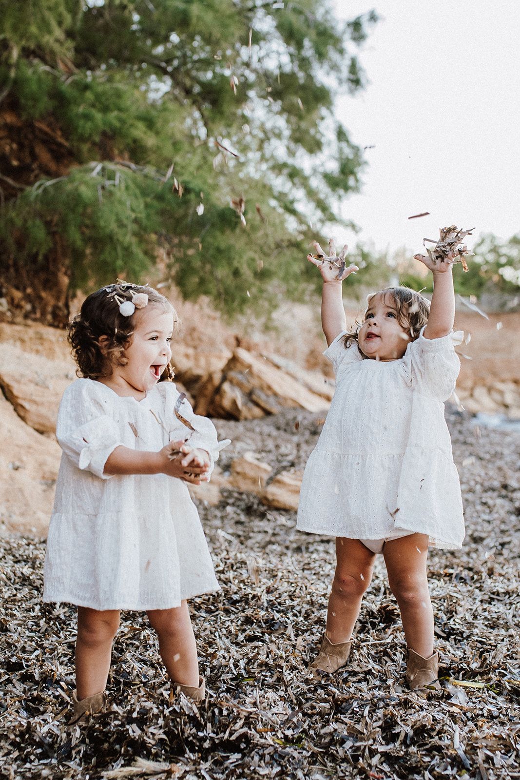 Two toddler girls in white dresses playing and tossing debris on a rocky beach.