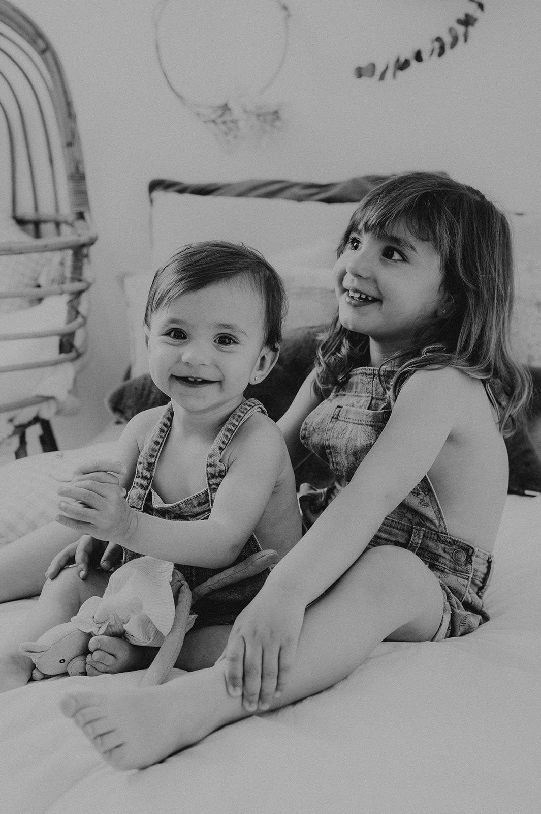 Two young siblings in matching denim overalls smile together on a bed in a black and white portrait.