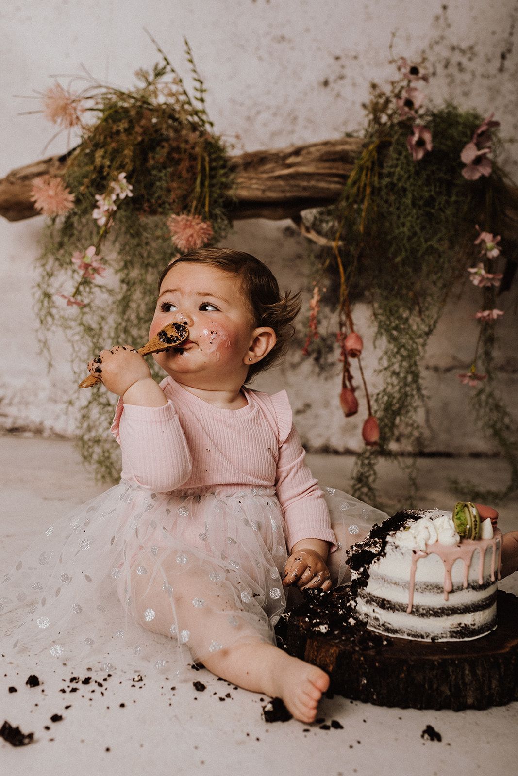 Baby girl in pink tutu smashing her first birthday cake, with frosting on her face, seated before floral backdrop.