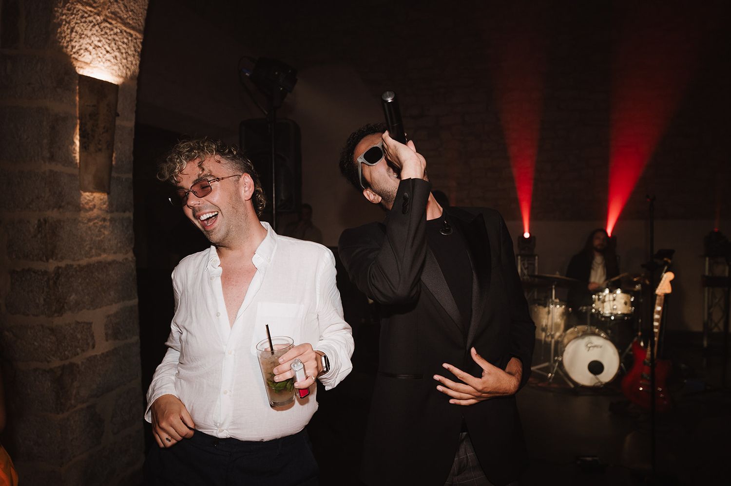 Two men laughing and dancing together at a wedding reception with red-lit stage and band in background.