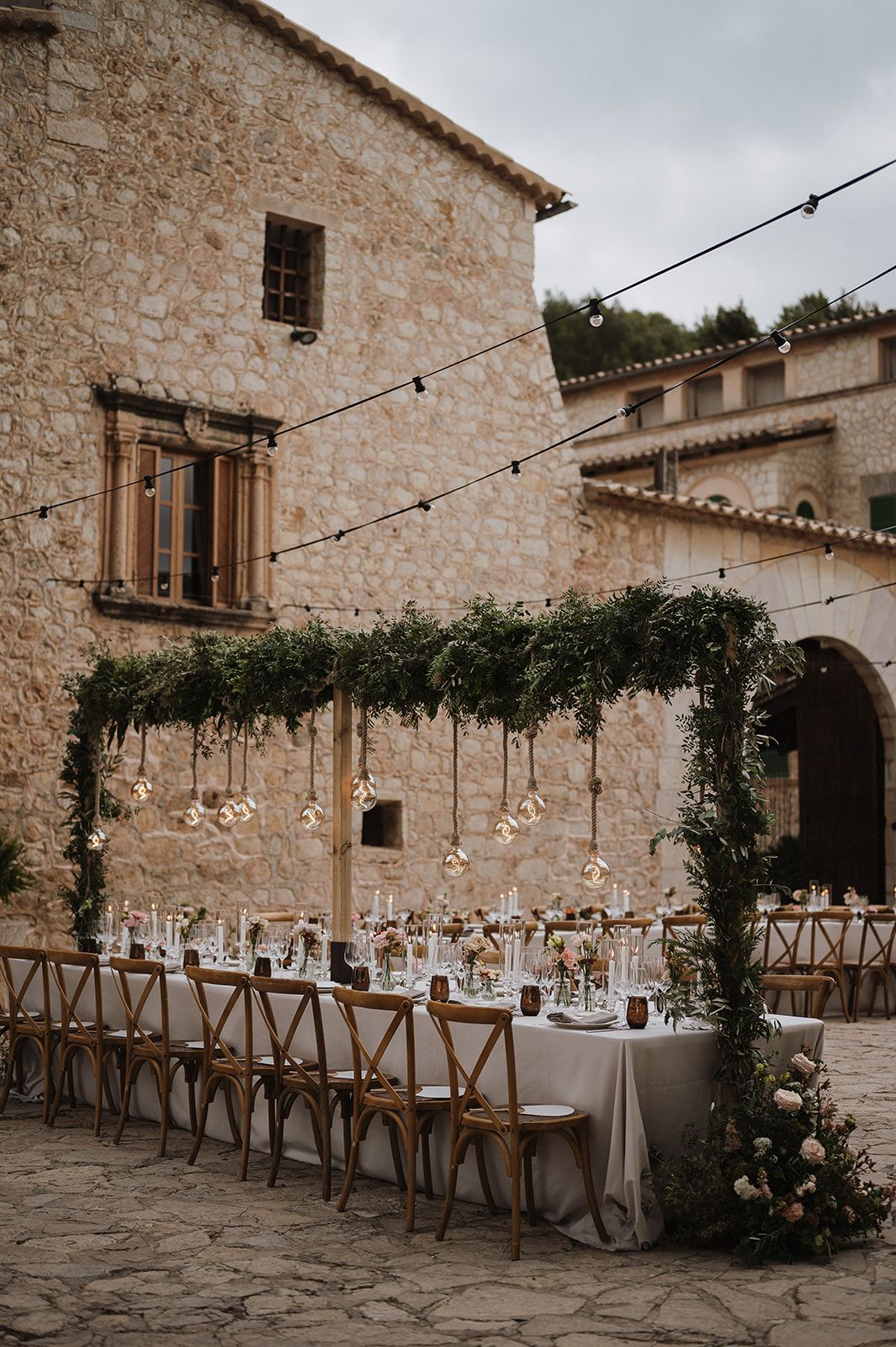 Elegant outdoor wedding reception at a rustic stone venue with long tables, greenery arch, and string lights.