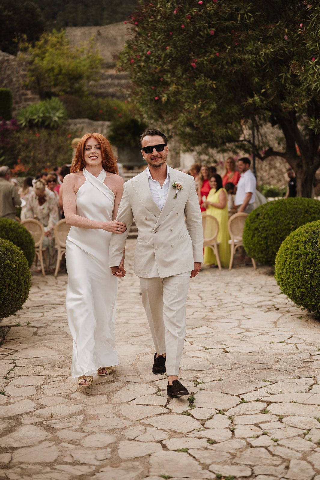 Radiant bride and groom walk down cobblestone aisle smiling after outdoor wedding ceremony in garden.