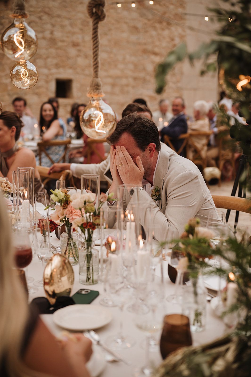 Emotional groom covering his face with hands during heartfelt wedding reception speeches at candlelit table.