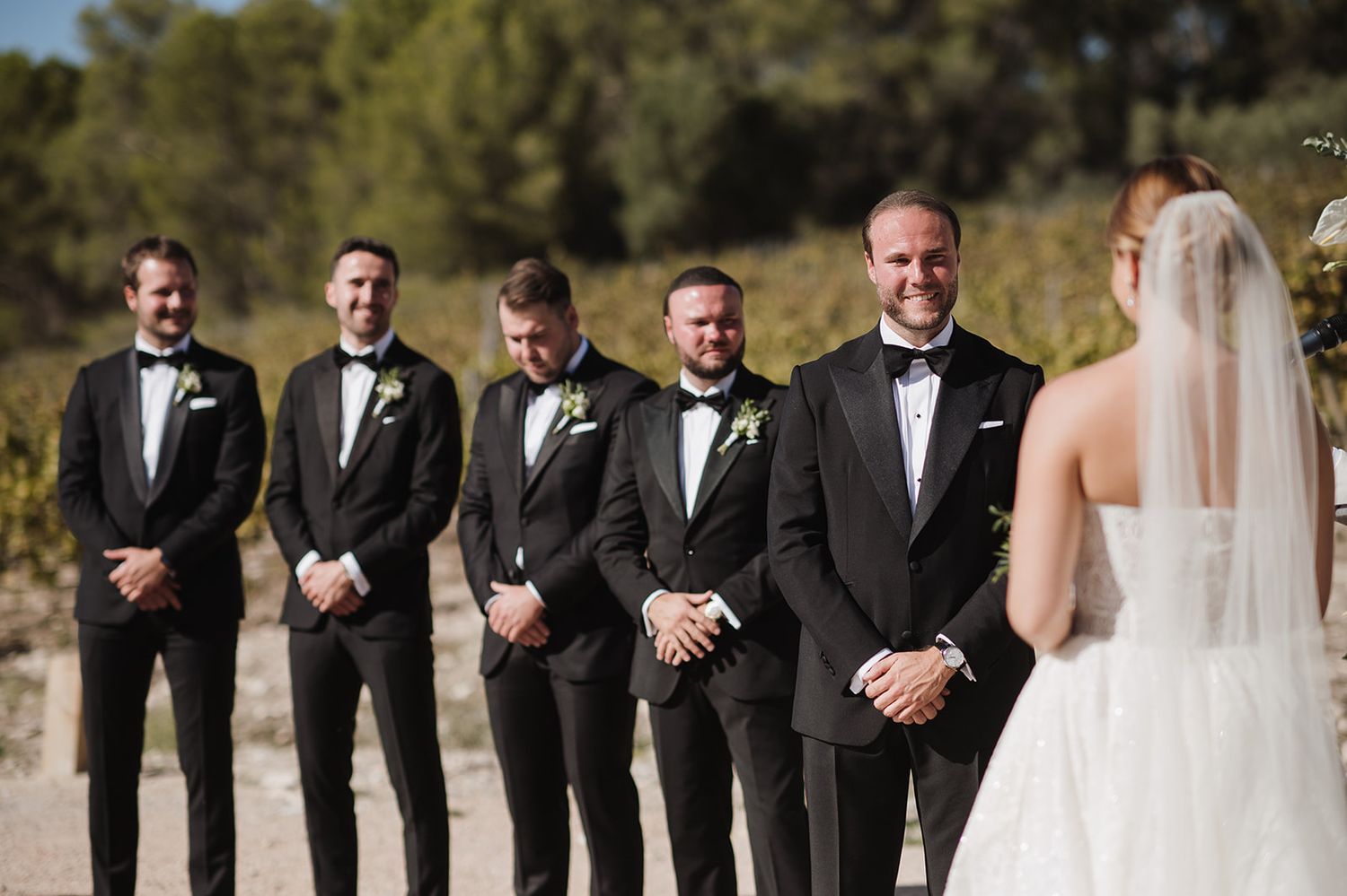 Groom smiling at bride approaching during outdoor wedding ceremony with groomsmen.