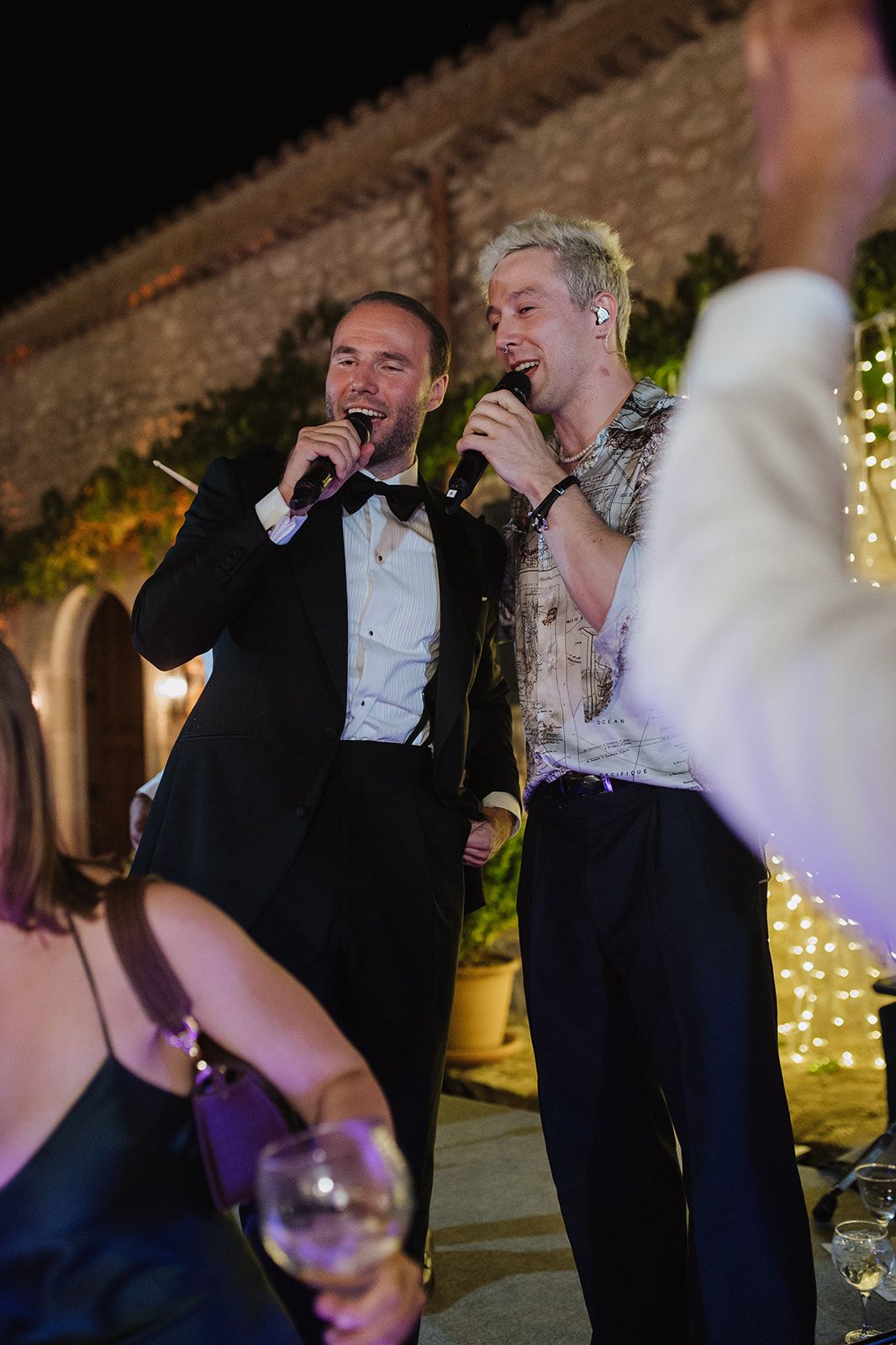 Groom and blonde performer singing into microphones together at lively outdoor wedding reception.