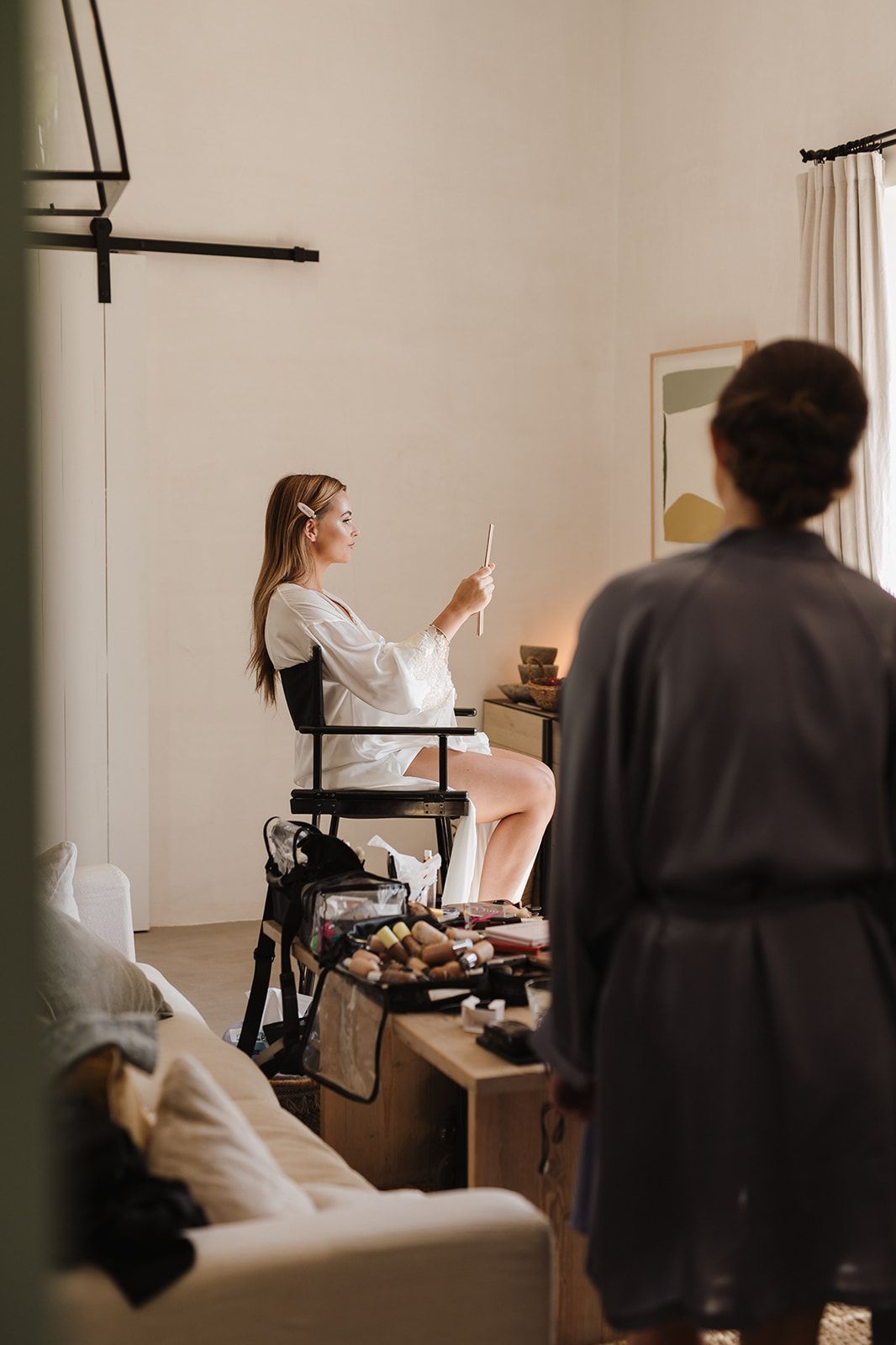 Bride in white robe sits in makeup chair checking phone while bridesmaid in grey robe stands nearby.