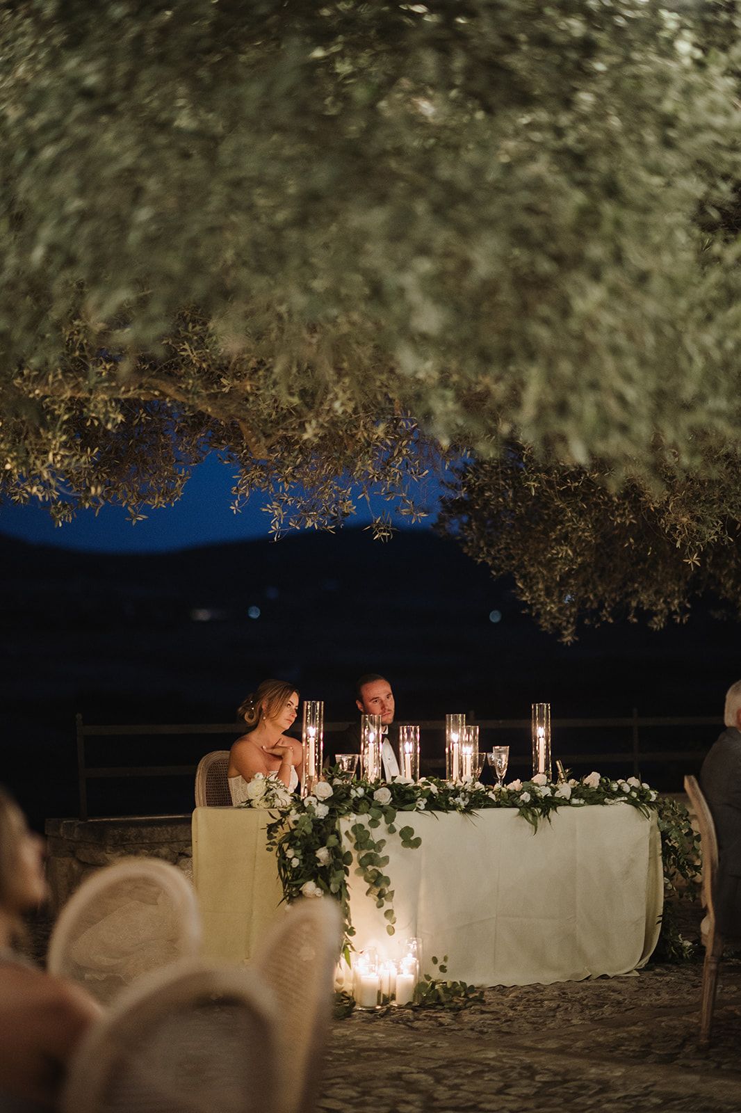 Bride and groom seated at candlelit sweetheart table beneath a tree during an elegant evening reception.