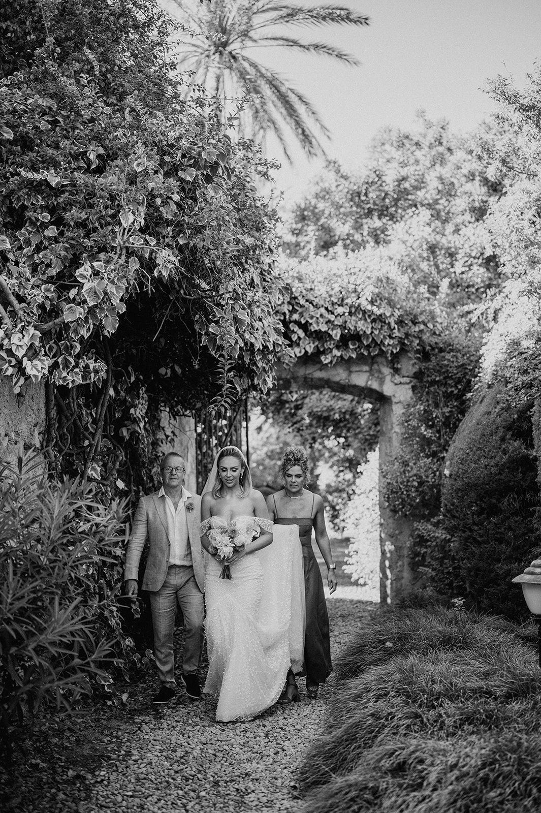 Bride walks through a lush garden archway with two companions during an outdoor wedding celebration.
