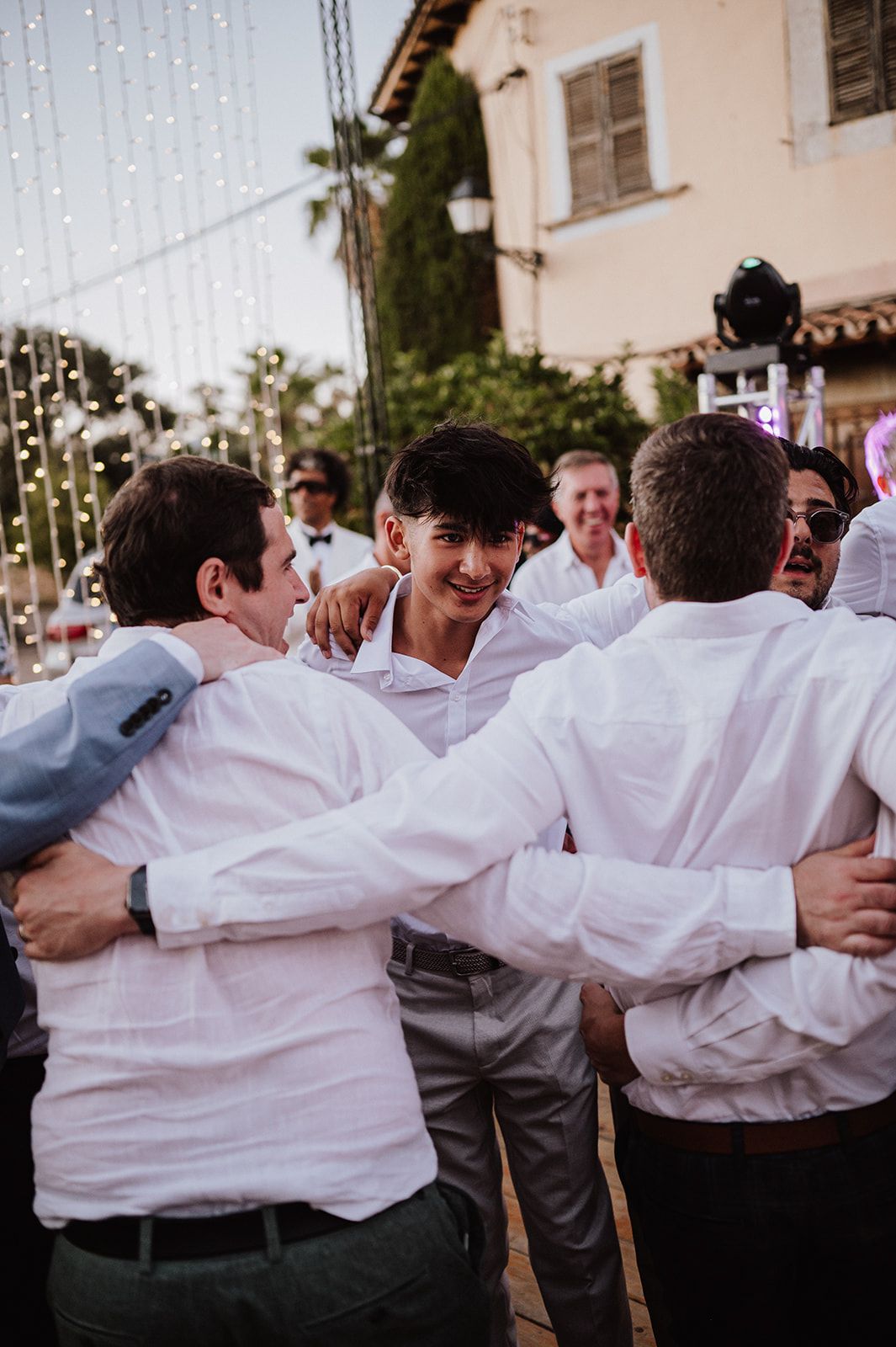 Groom embraces group of men in white shirts during festive outdoor wedding reception.
