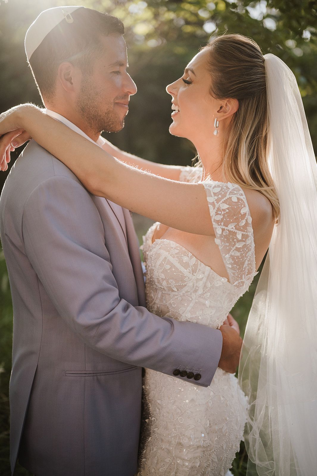 Bride in beaded lace gown with veil embraces groom in grey suit during golden hour portraits outdoors.