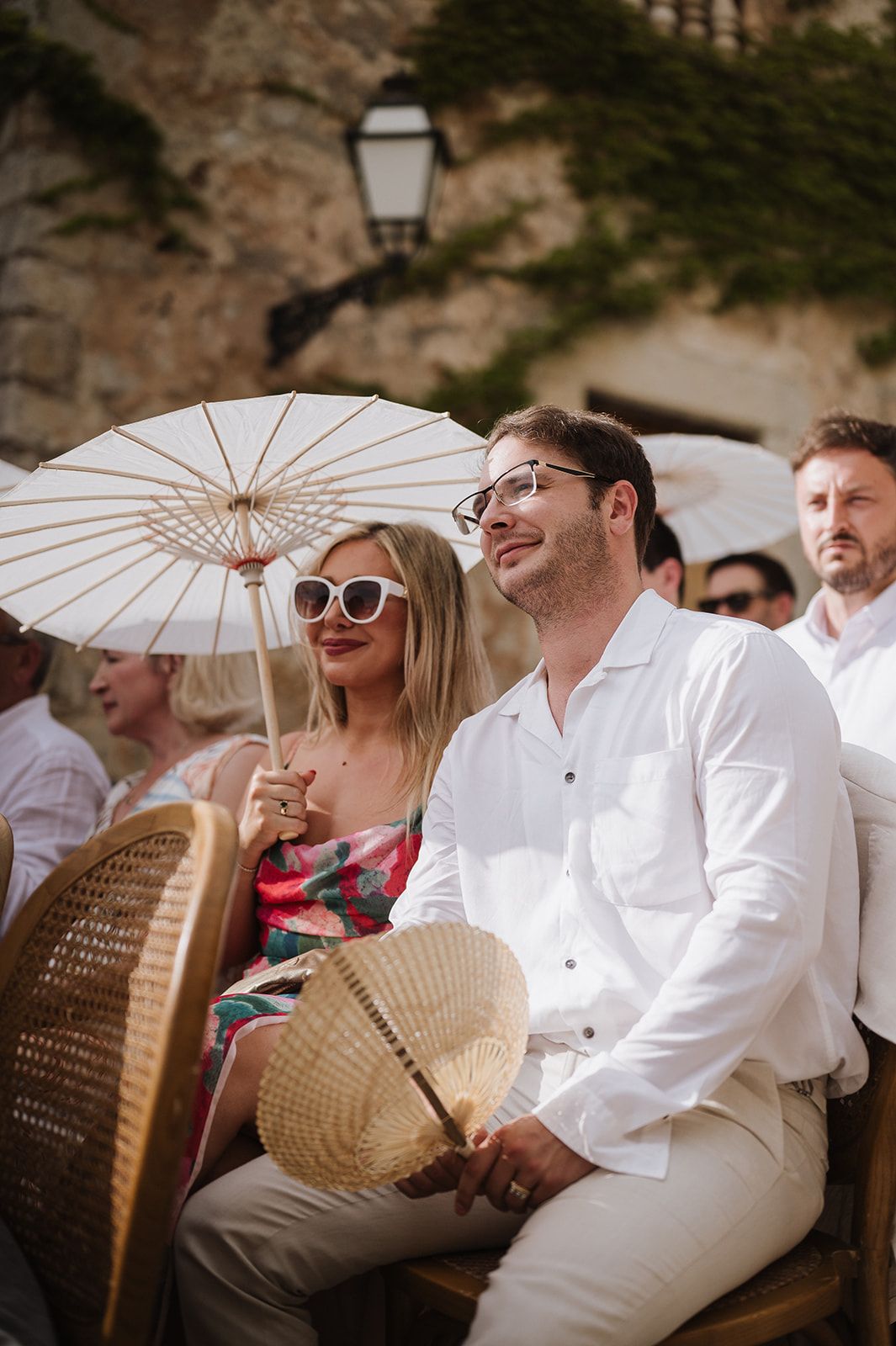 Wedding guests seated outdoors, woman in floral dress holding white parasol beside man in white shirt at sunny ceremony.