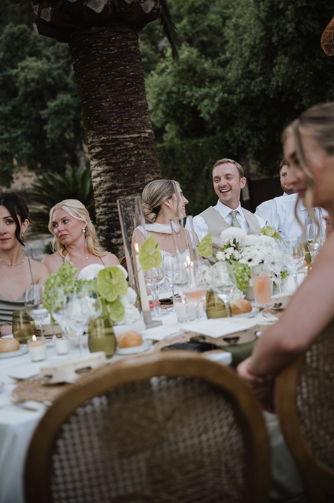 Wedding guests laugh and smile at elegant outdoor reception table adorned with white floral centerpieces.