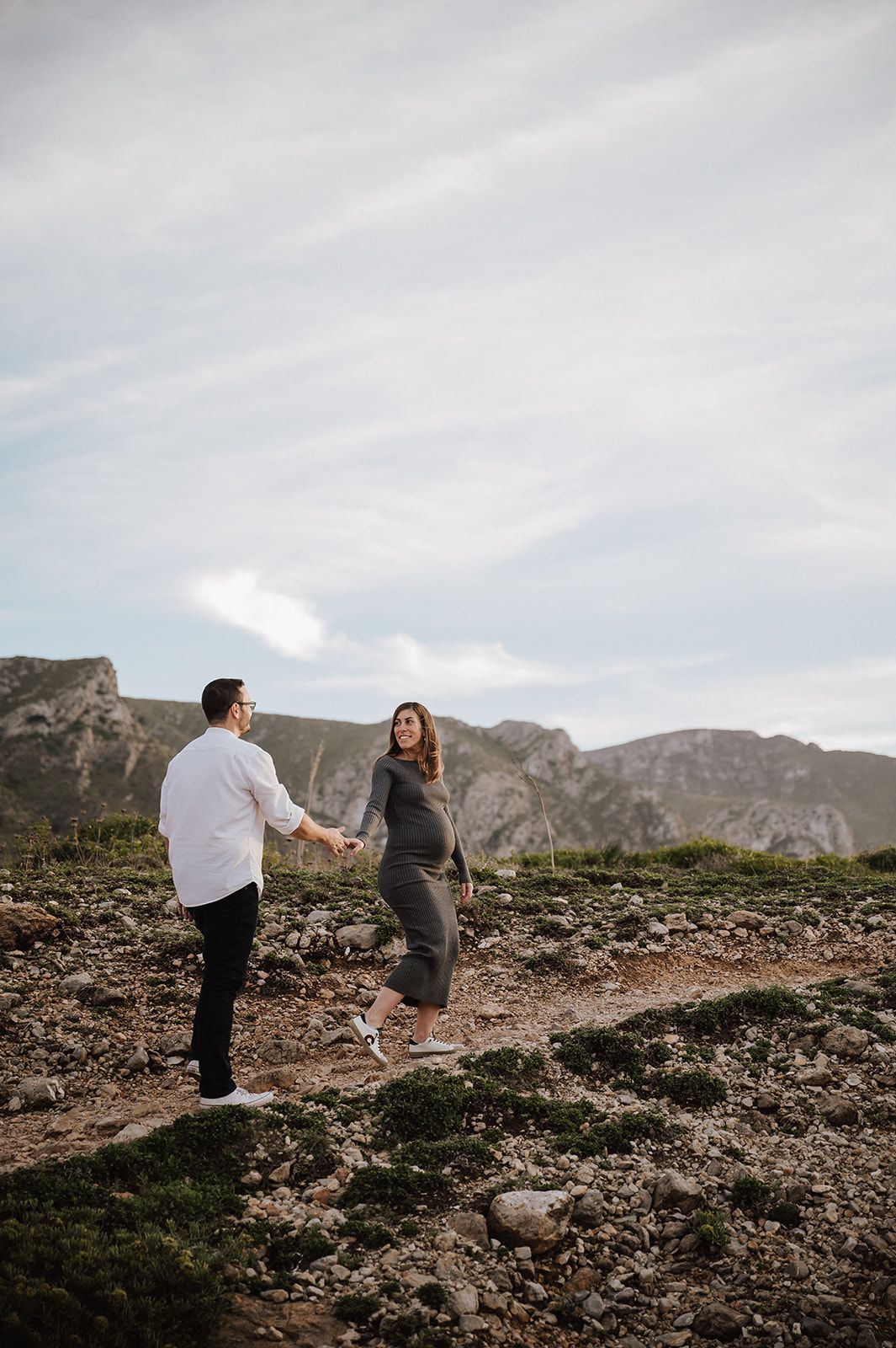 Couple holding hands on rocky mountain terrain, pregnant woman in gray dress with man in white shirt, cloudy sky backdrop.