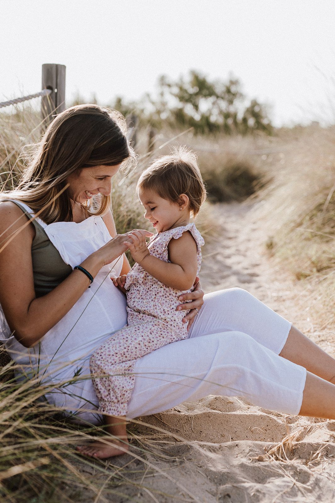 A mother and toddler sit together on sandy beach dunes, sharing a tender moment in warm golden sunlight.