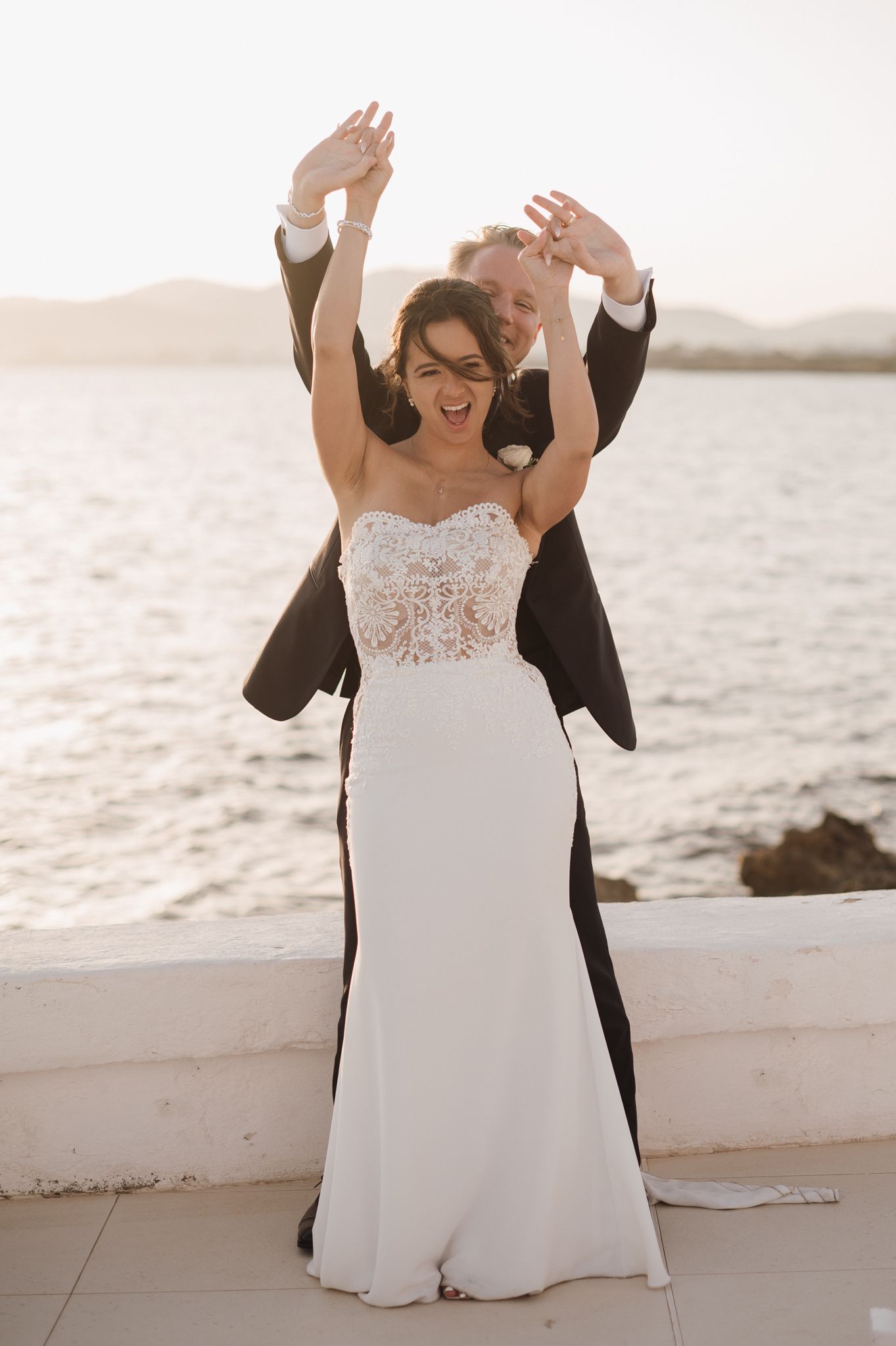 Joyful bride and groom celebrating on waterfront at sunset, arms raised, bride in lace wedding gown.