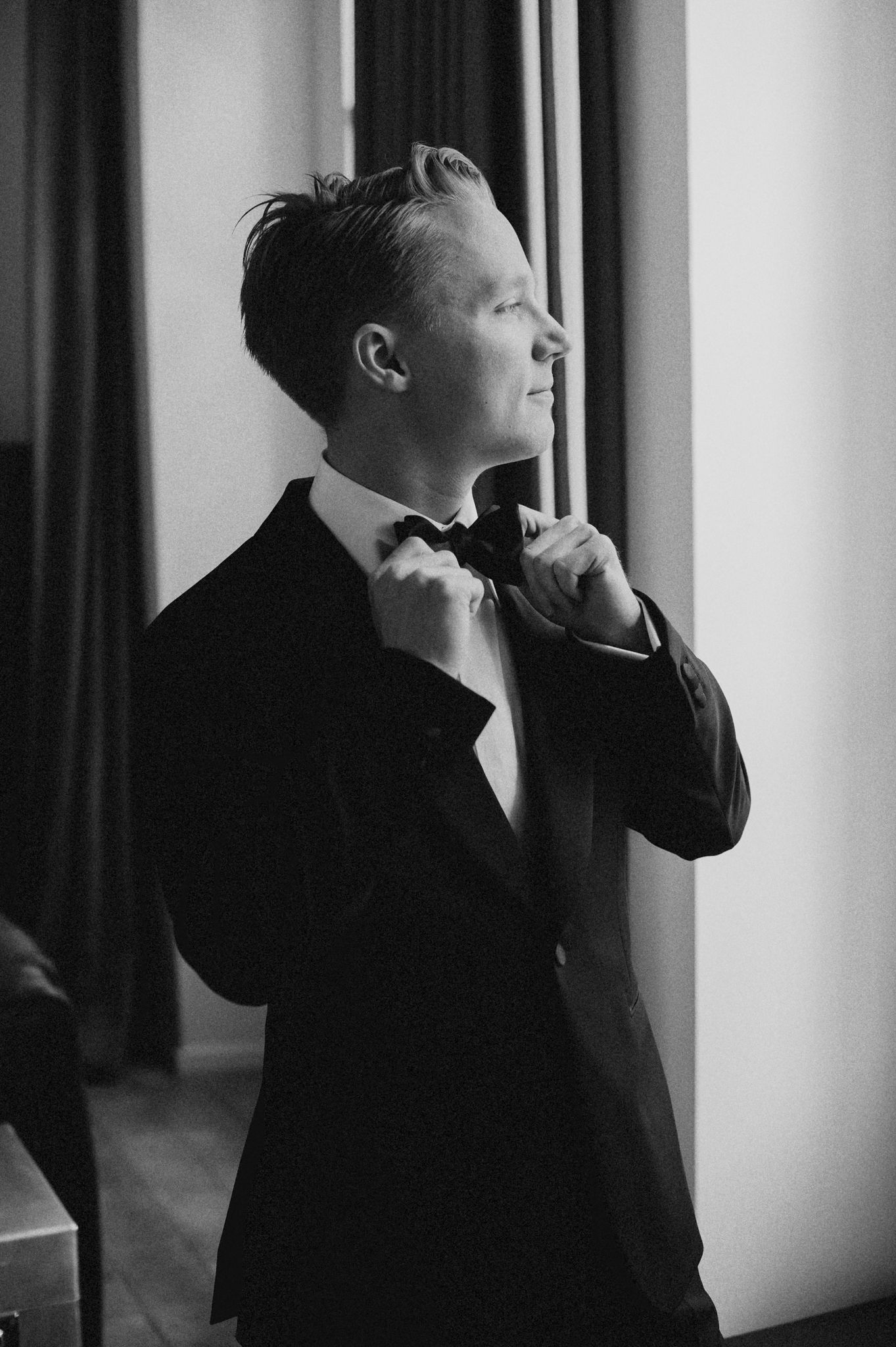 Black and white photo of a groom in a tuxedo adjusting his bow tie while standing near a window.