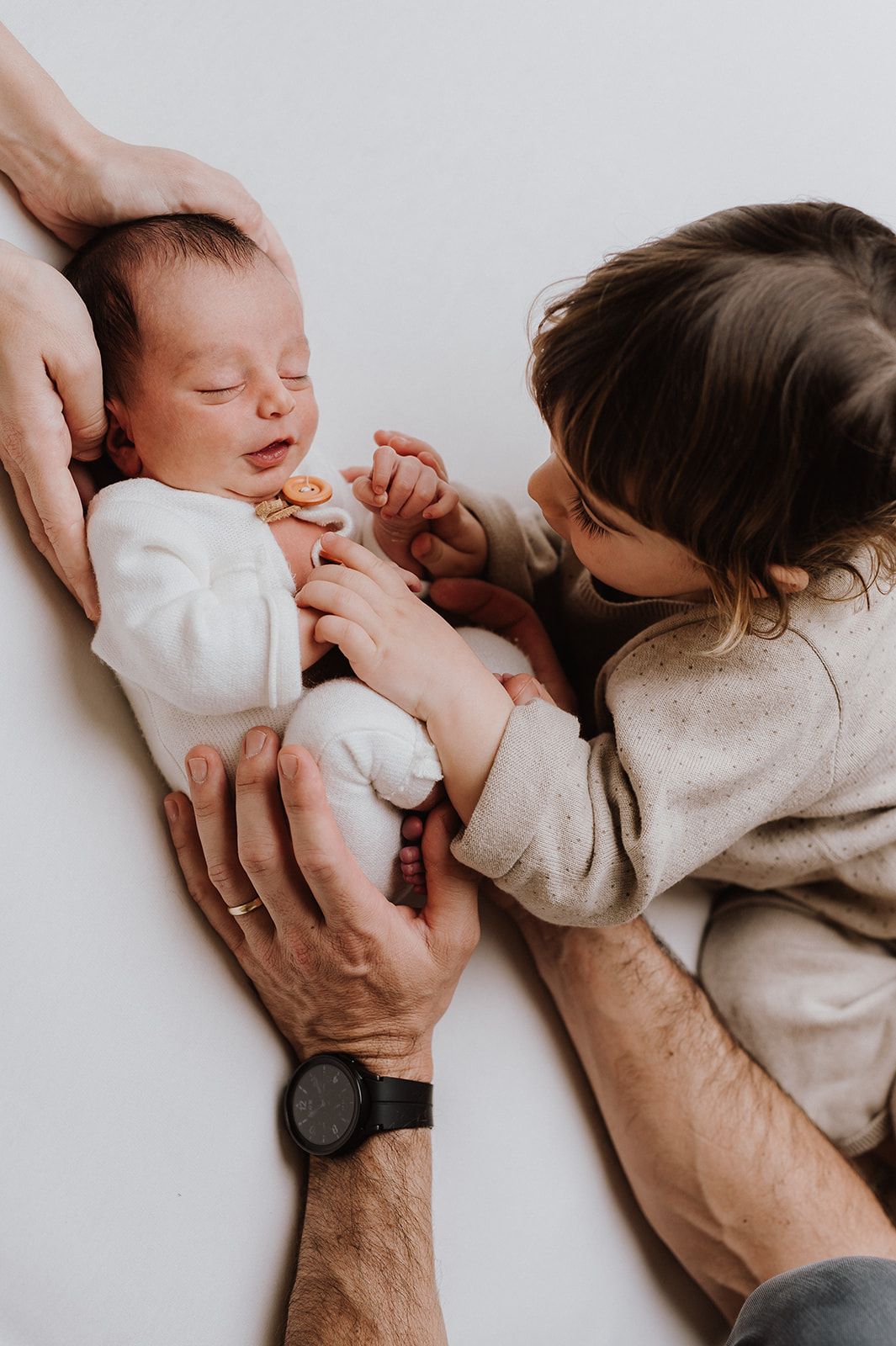A newborn baby in white onesie being held by parents while an older sibling leans in to give a gentle kiss.