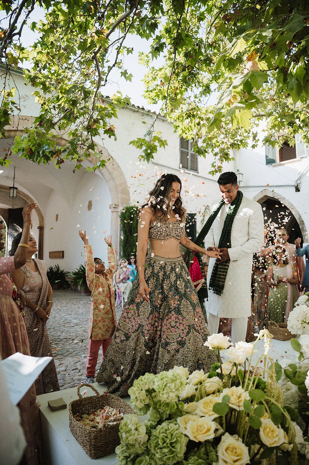 Bride in green embroidered lehenga and groom in white sherwani celebrate with confetti in a courtyard.