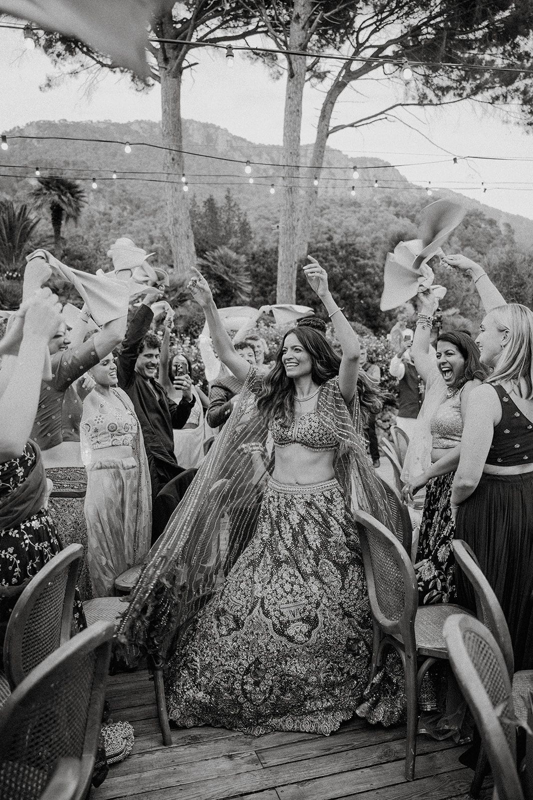 Black and white photo of bride in lehenga dancing joyfully surrounded by celebrating wedding guests.
