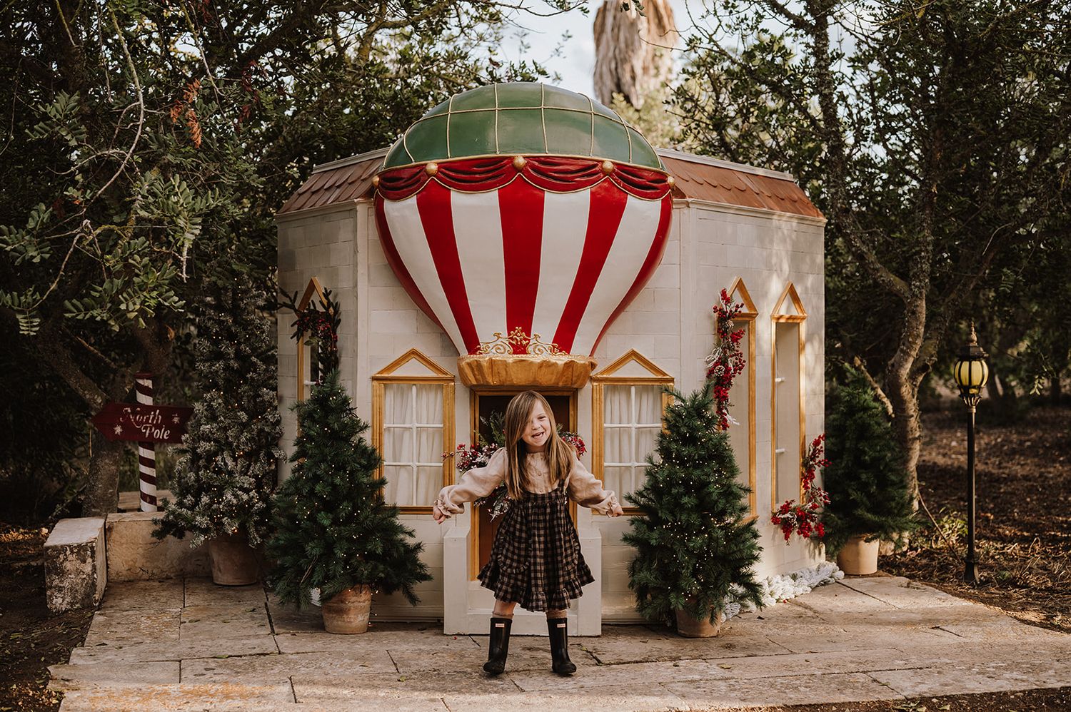 Girl poses before a whimsical hot air balloon-themed playhouse with Christmas trees and festive decor.