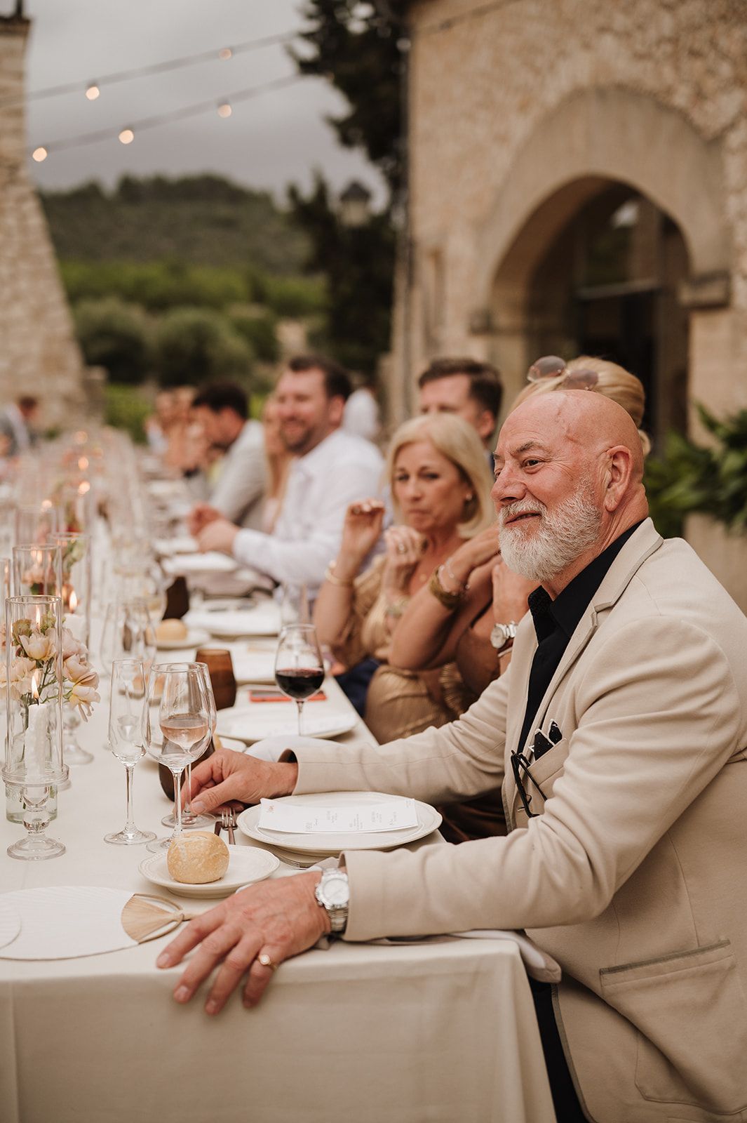 Elegant older gentleman in beige suit smiles at a long outdoor wedding reception table with wine glasses.