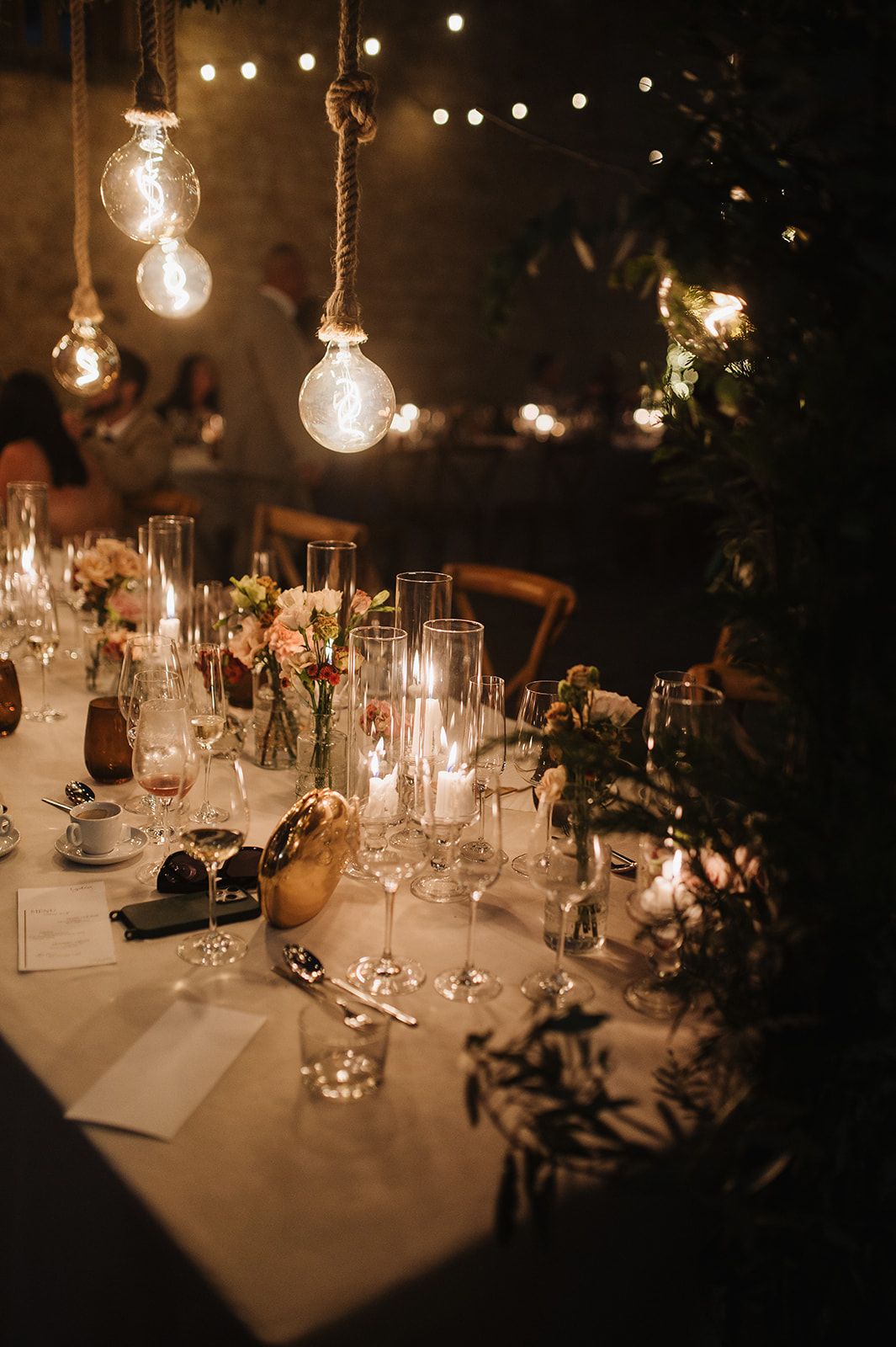 Romantic wedding table setting with hanging Edison bulb lights, candles, and floral centerpieces.