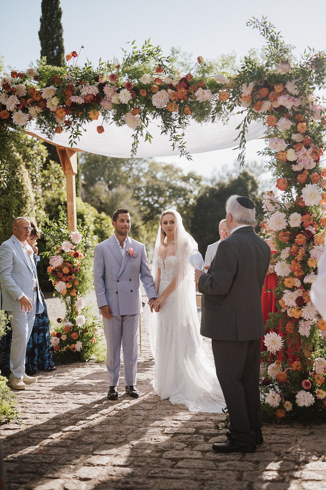 Couple exchanging vows under floral chuppah adorned with pink and orange blooms.
