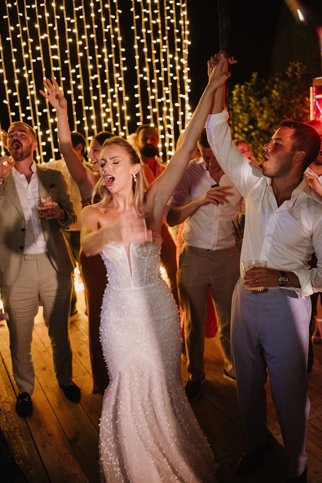 Bride in sequin gown dances with raised arms among guests at night wedding reception.