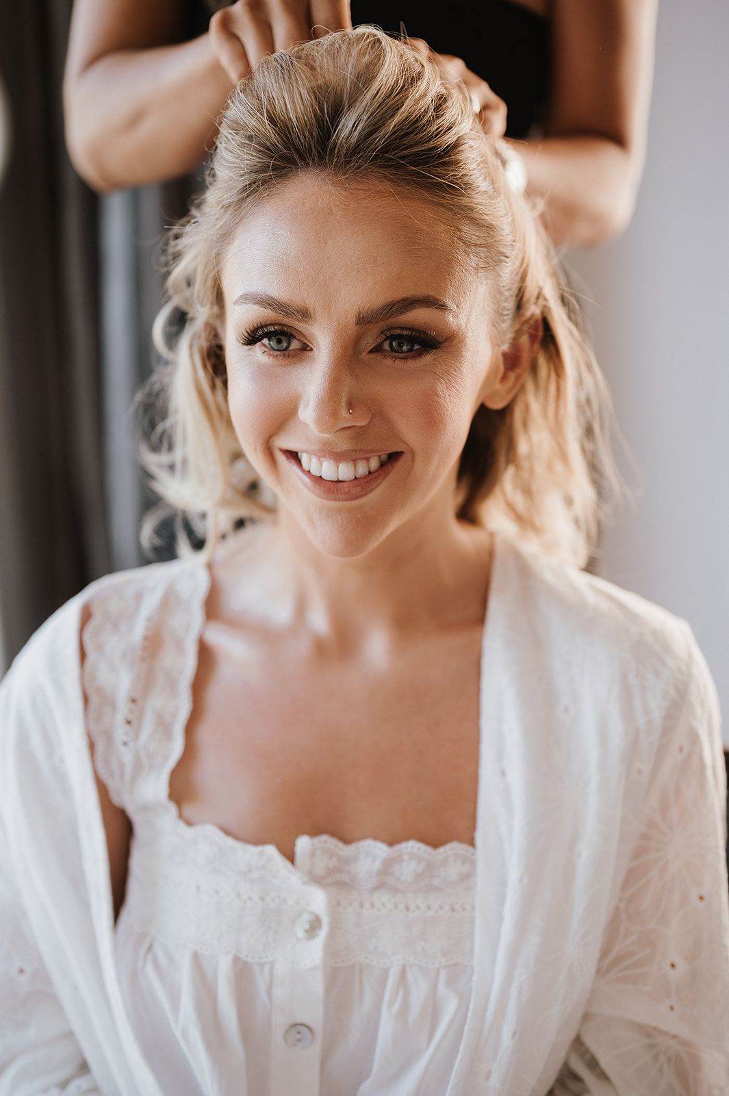 Smiling blonde bride in white robe getting her hair styled, with soft natural makeup and an elegant updo in progress.