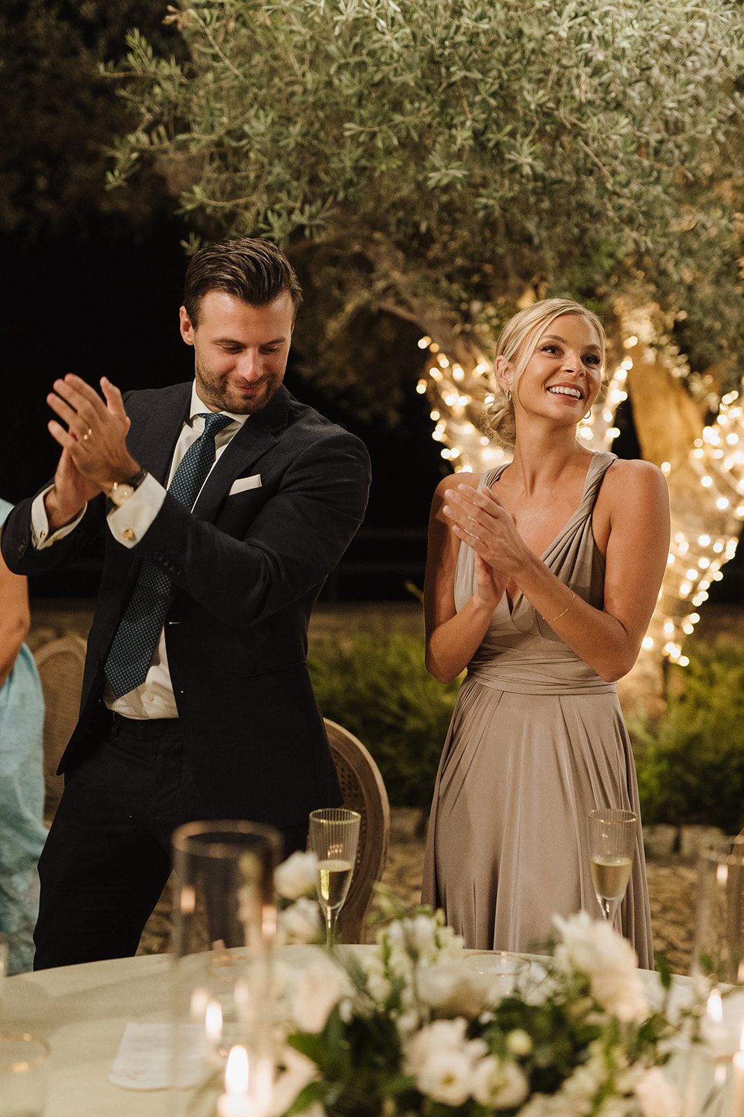 Smiling wedding guests in formal attire applauding at elegant outdoor candlelit wedding reception.