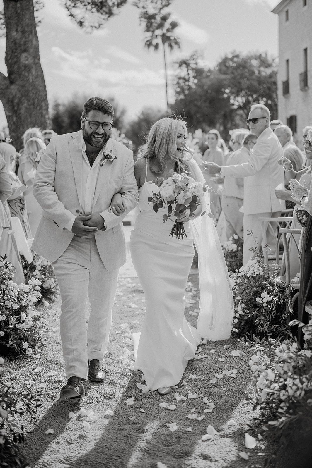Joyful newlyweds walk back down the aisle as guests toss flower petals at outdoor wedding.