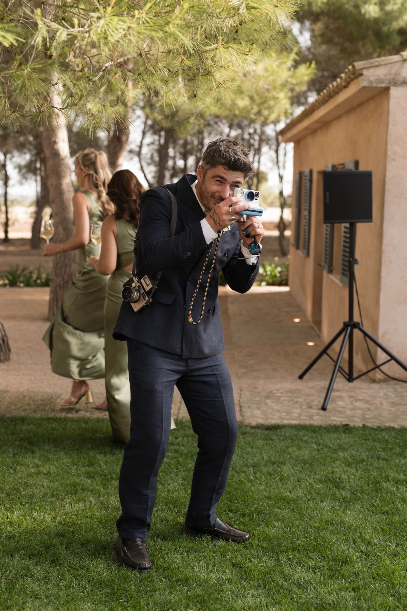 Wedding guest in navy suit playfully posing with camera during outdoor wedding reception on green lawn.