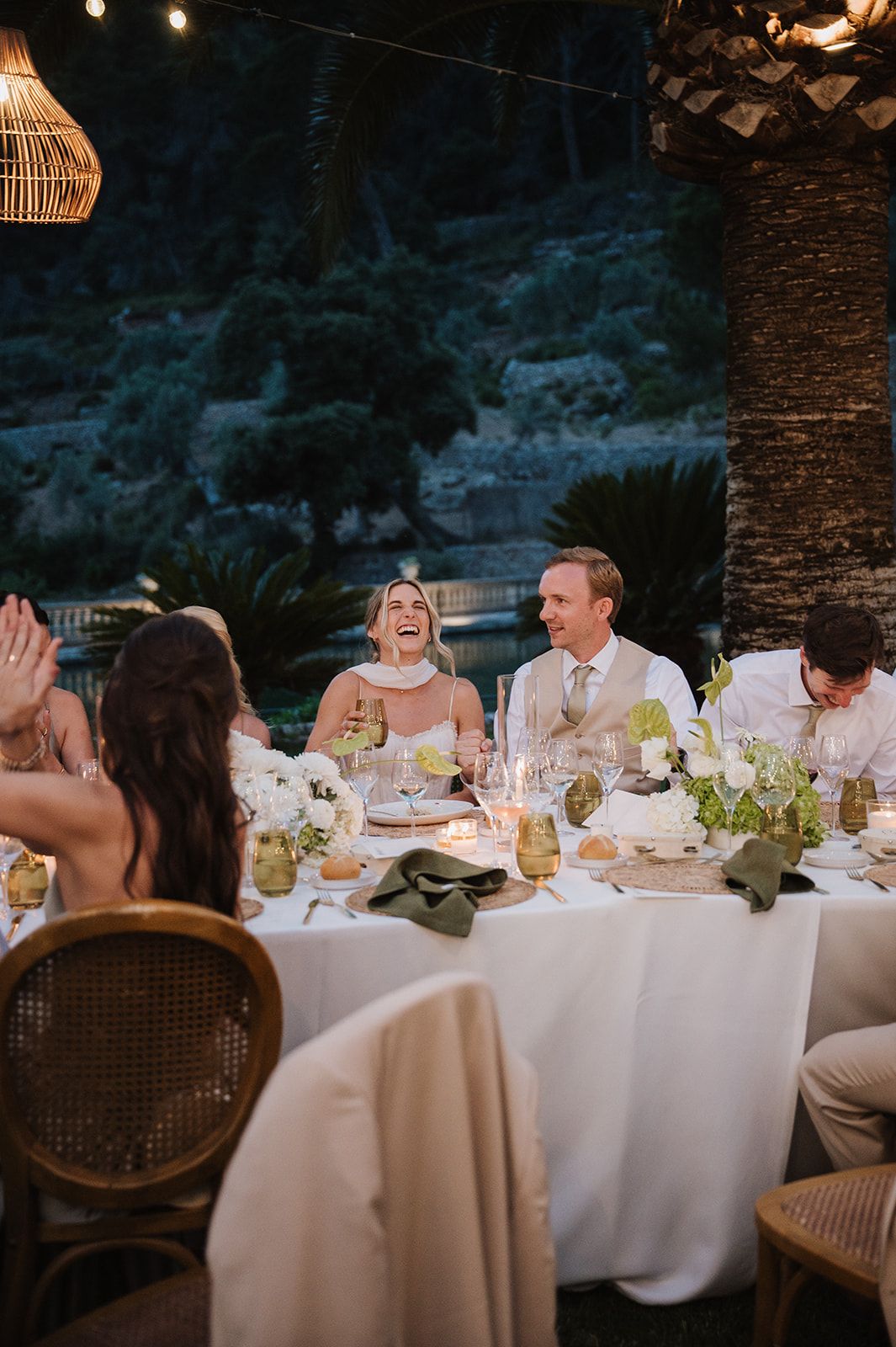 Bride and groom laughing joyfully at candlelit outdoor wedding reception dinner table surrounded by guests at night.