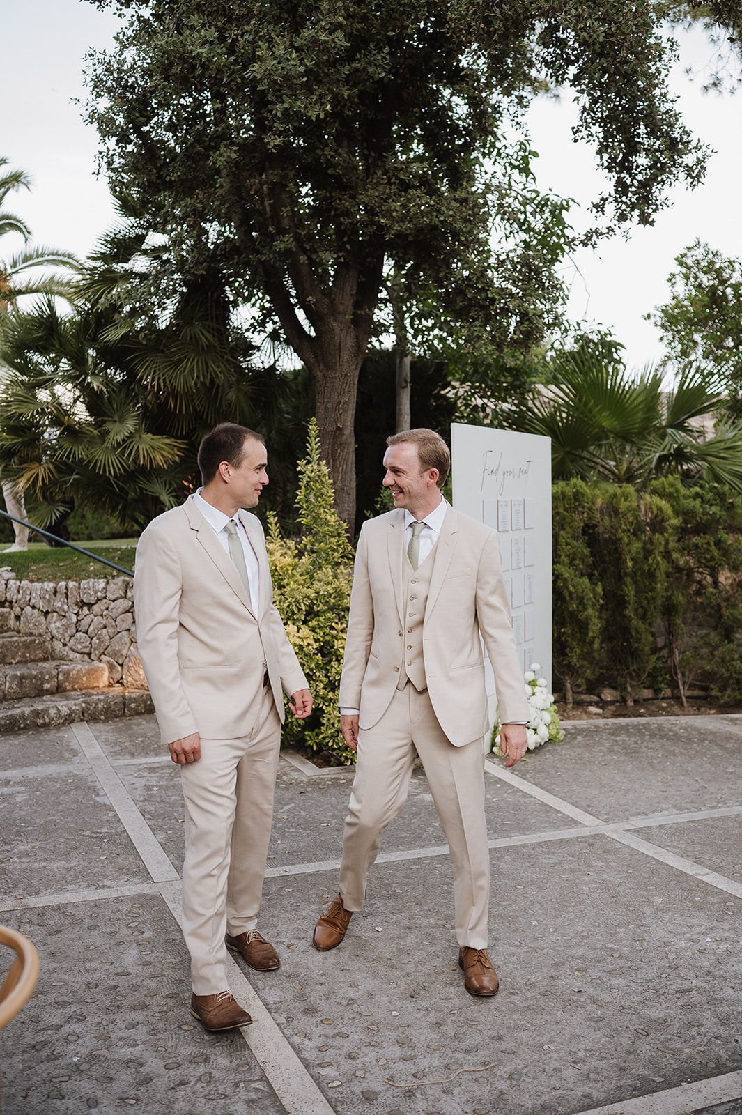 Two groomsmen in matching beige suits share a smile while standing on a sunlit outdoor patio.