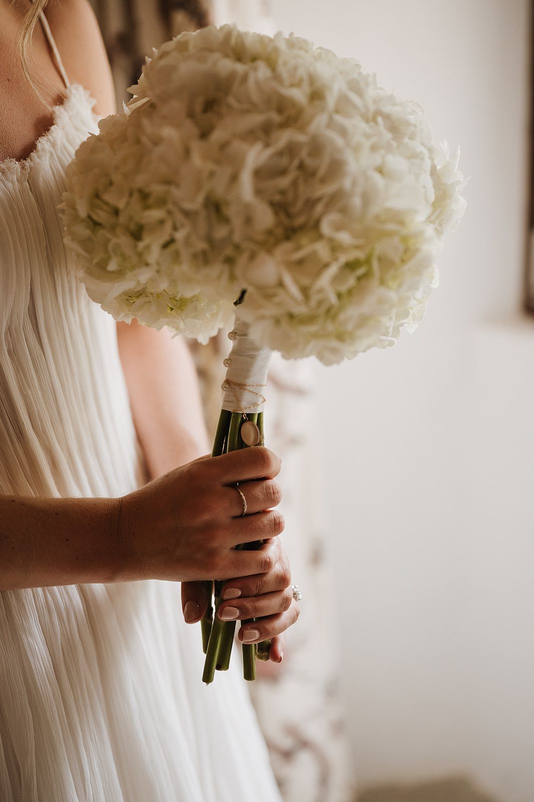 Bride in white pleated gown holding a lush white hydrangea bouquet wrapped with ribbon on her wedding day.