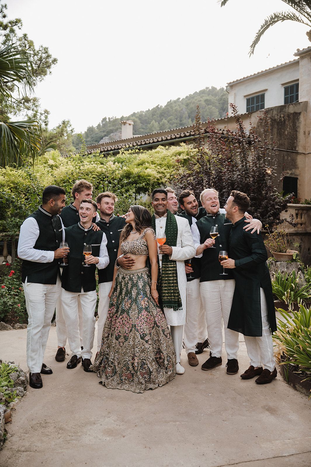 Groom in embroidered lehenga posing with groomsmen in dark vests at Indian wedding.
