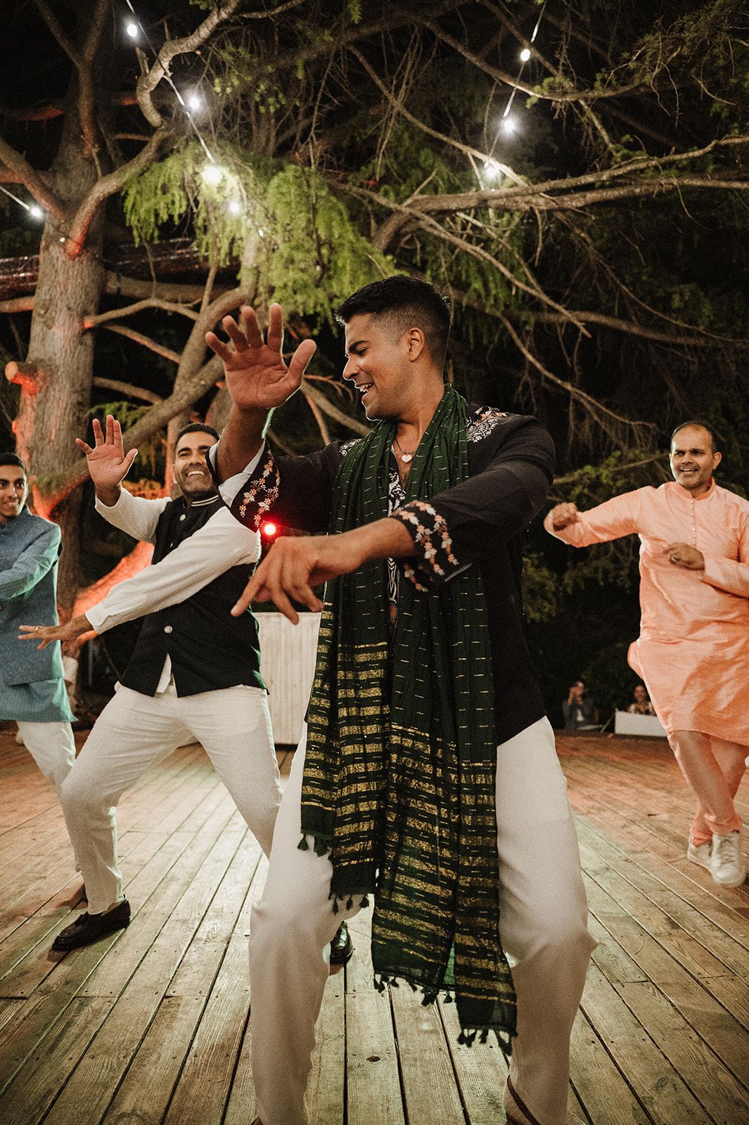 Groom in black outfit with green dupatta dancing energetically with guests at outdoor Indian wedding reception.