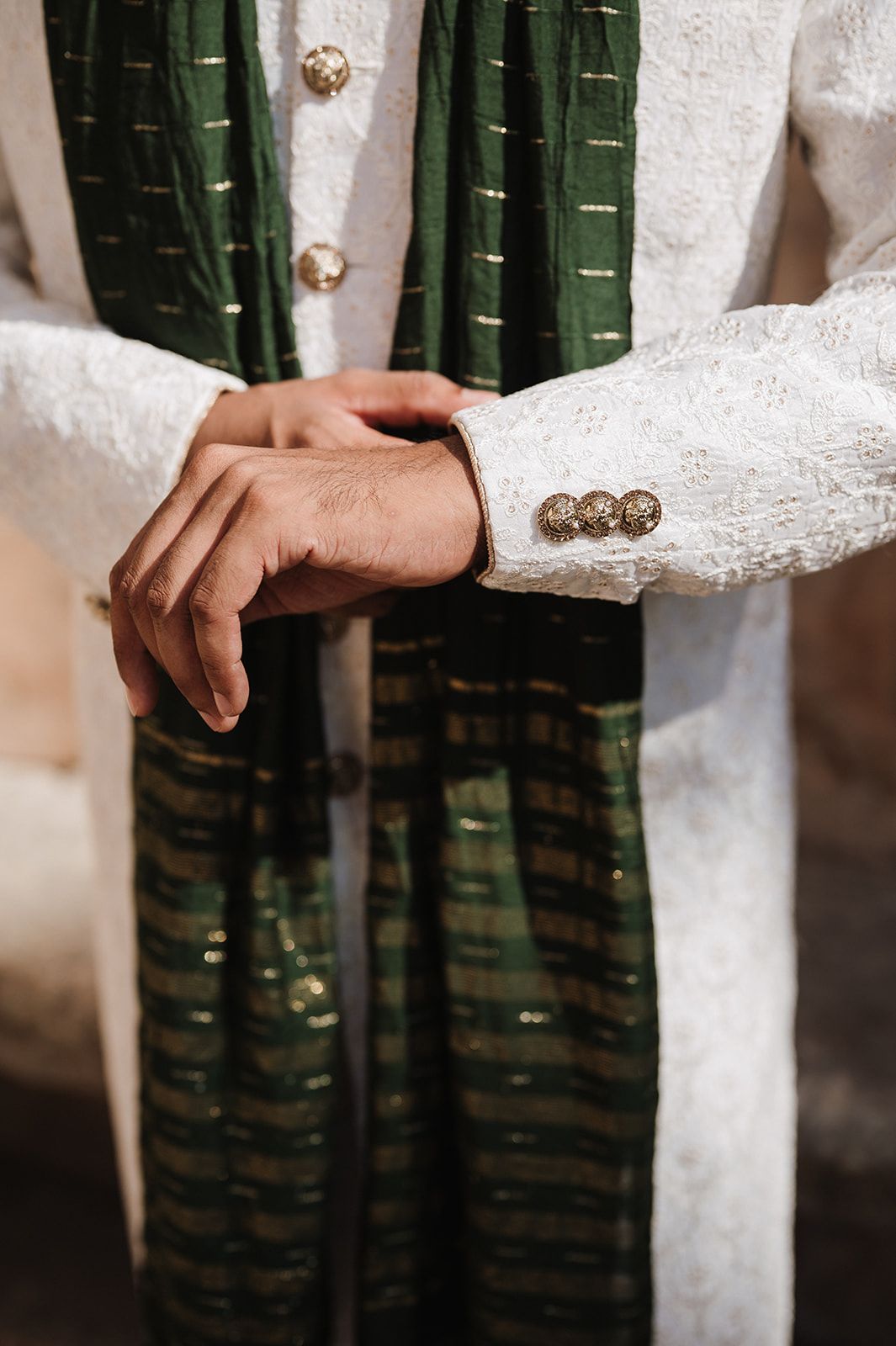 Close-up of groom in white sherwani with gold buttons and green embroidered dupatta at Indian wedding.