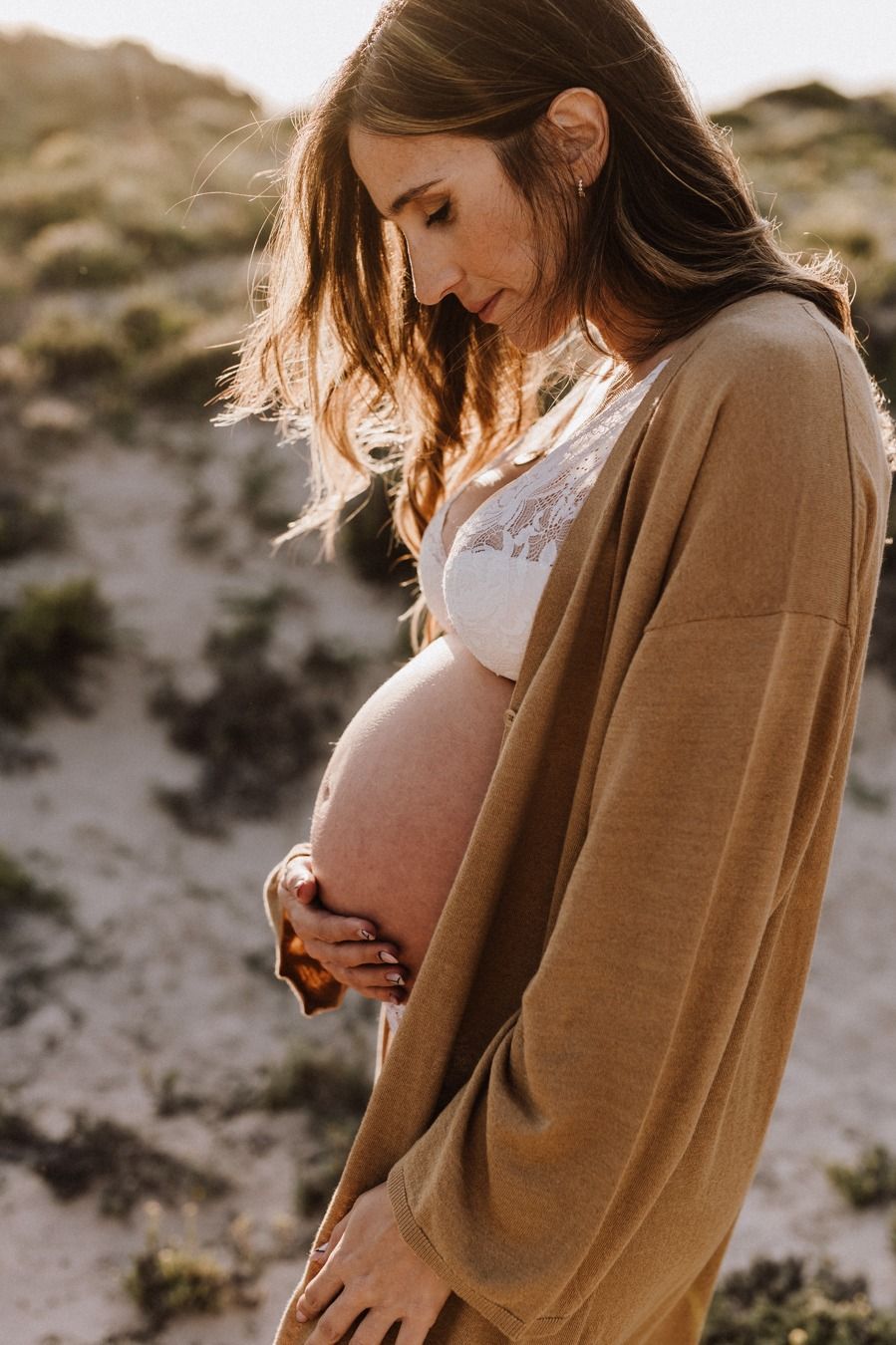 Pregnant woman in tan cardigan and white lace bra cradling her baby bump outdoors at golden hour.