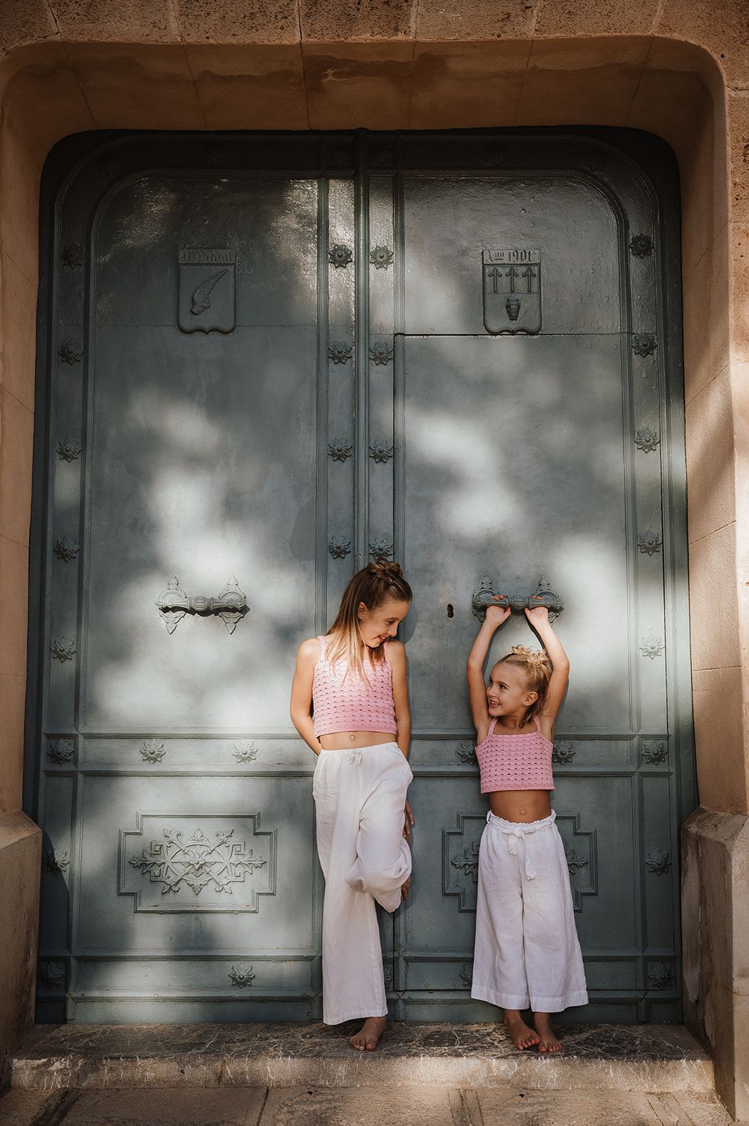 Two girls in matching pink crop tops and white wide-leg pants pose in front of a large ornate green door.
