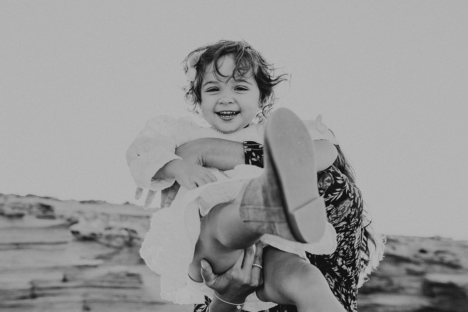 Black and white photo of a laughing toddler being lifted up outdoors on a beach.