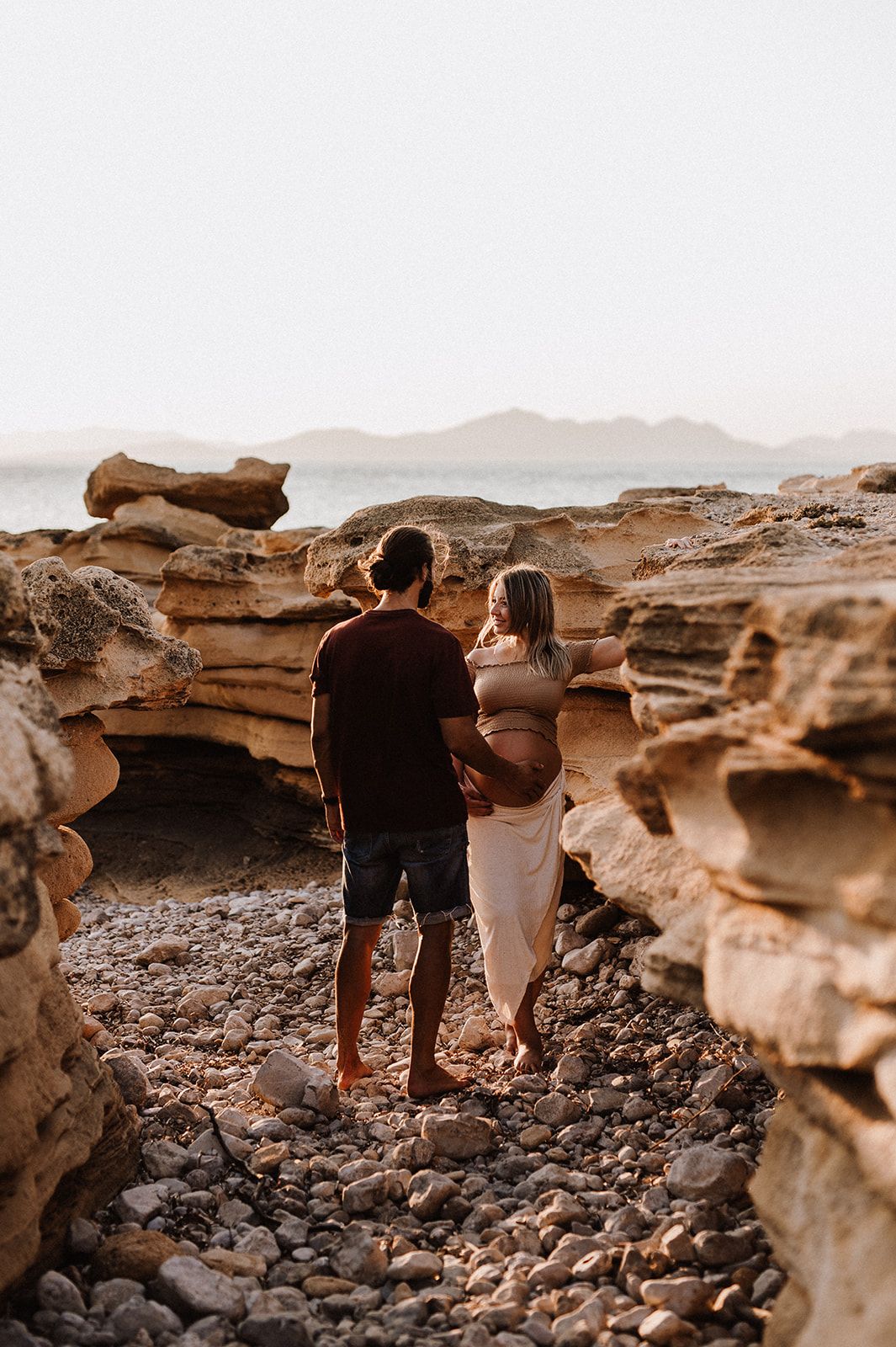 Pregnant couple standing among rocky coastal landscape, embracing while overlooking calm ocean at sunset.