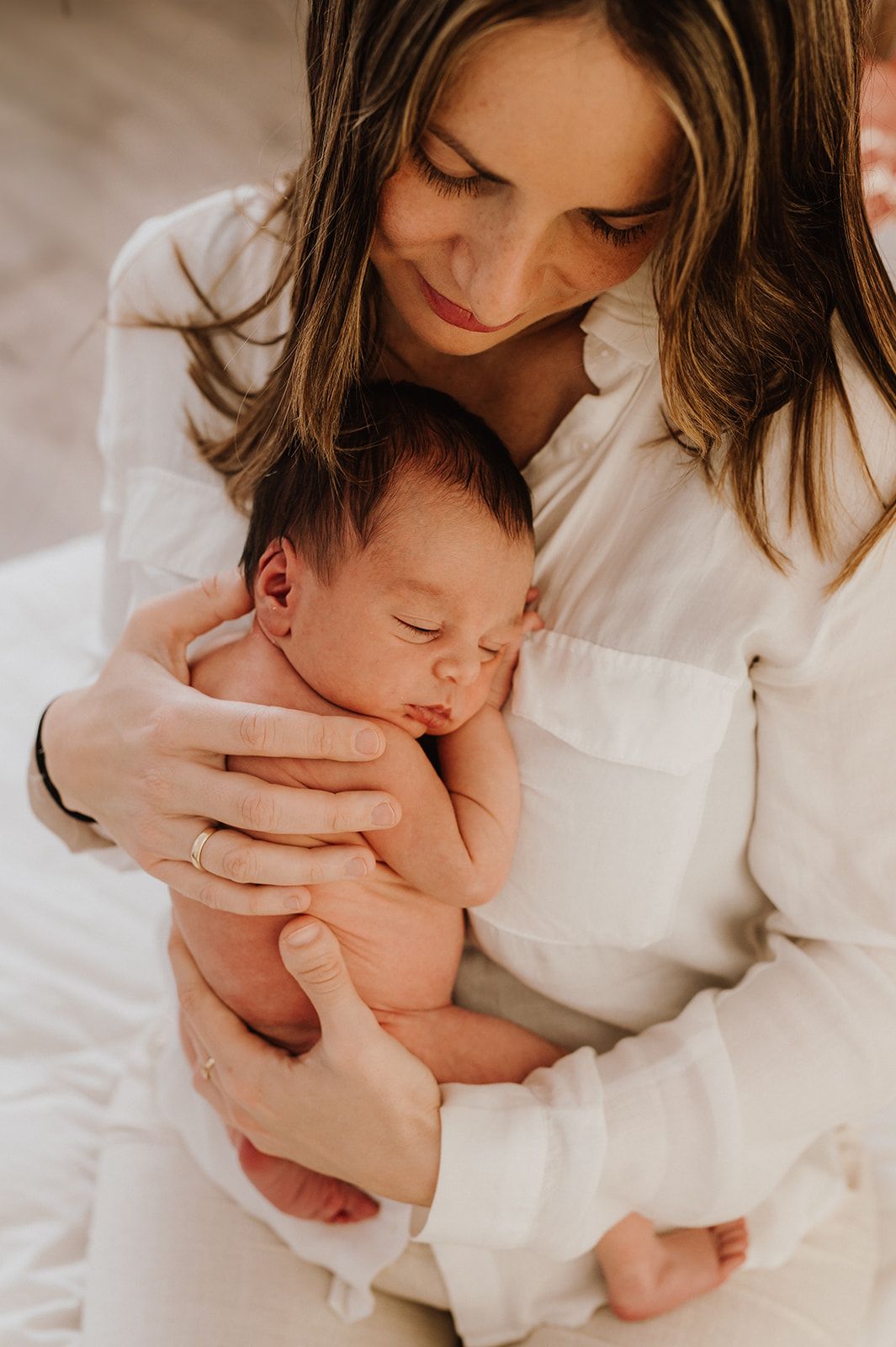 Mother in white shirt tenderly holding sleeping newborn baby close to her chest.
