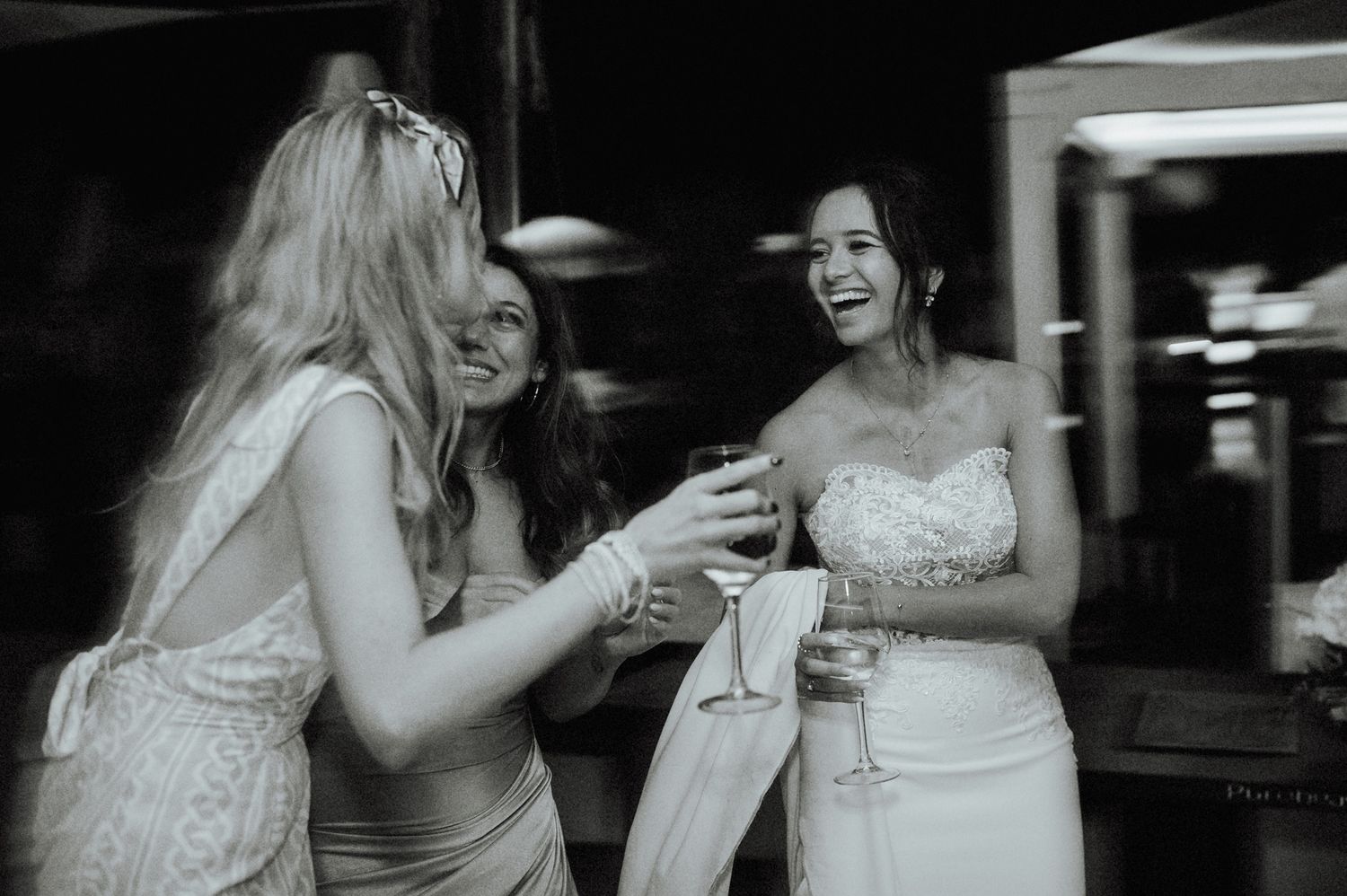 Black and white candid photo of laughing bride in strapless lace gown celebrating with blonde female guest.
