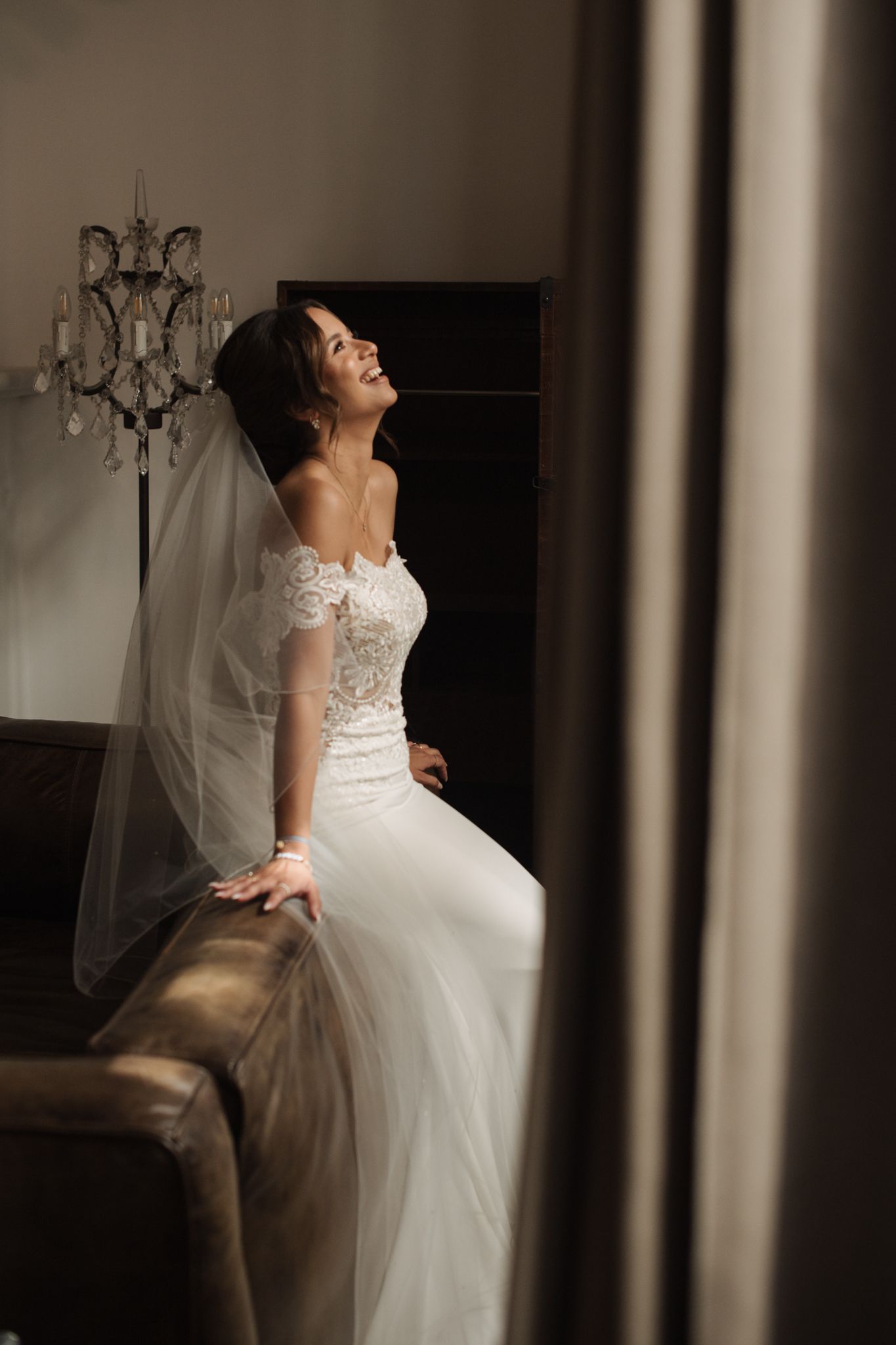 Radiant bride in lace off-shoulder wedding gown and veil seated on sofa laughing joyfully in elegant room.