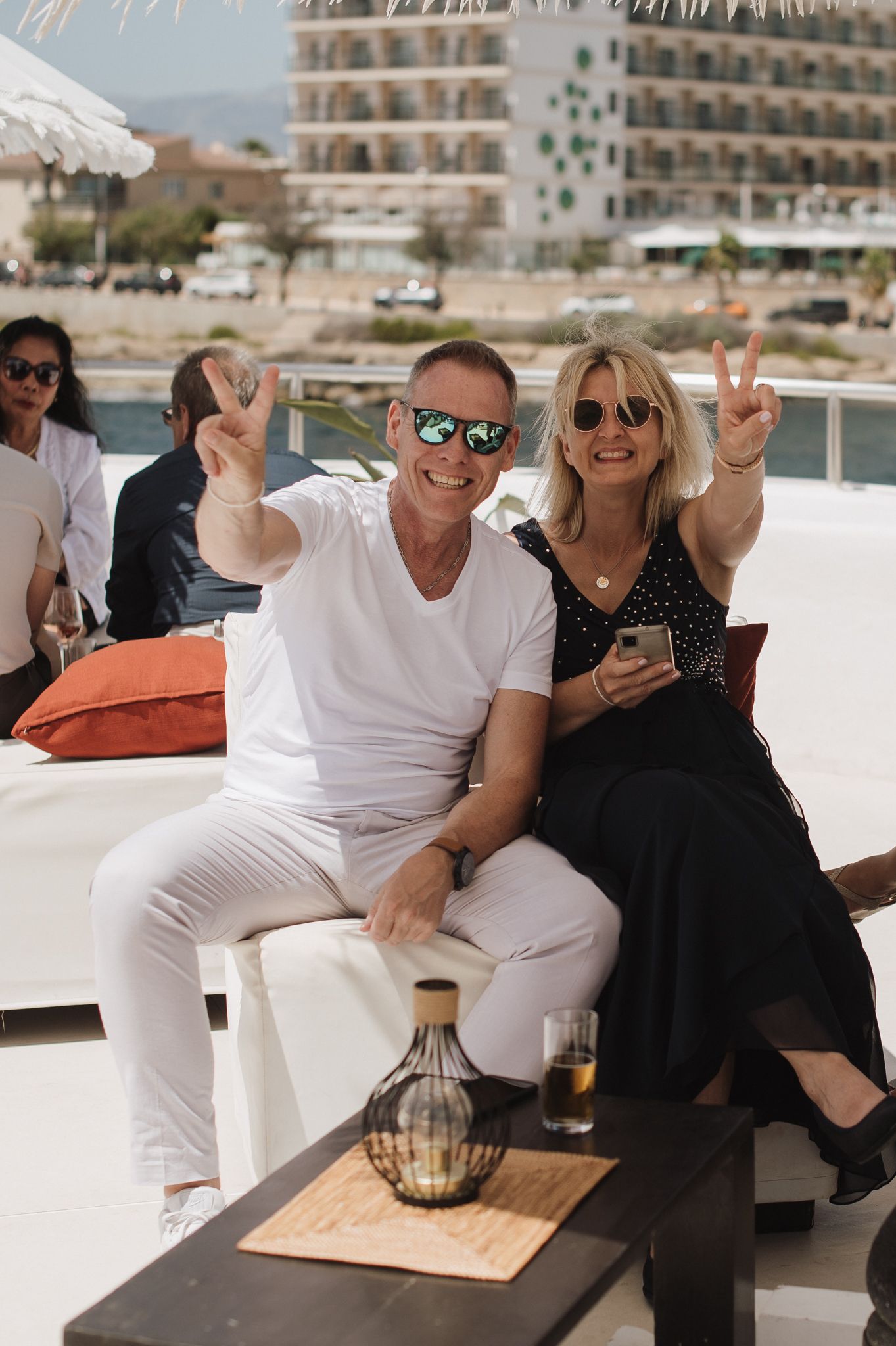 Smiling couple in sunglasses pose with peace signs at sunny outdoor wedding reception by the sea.